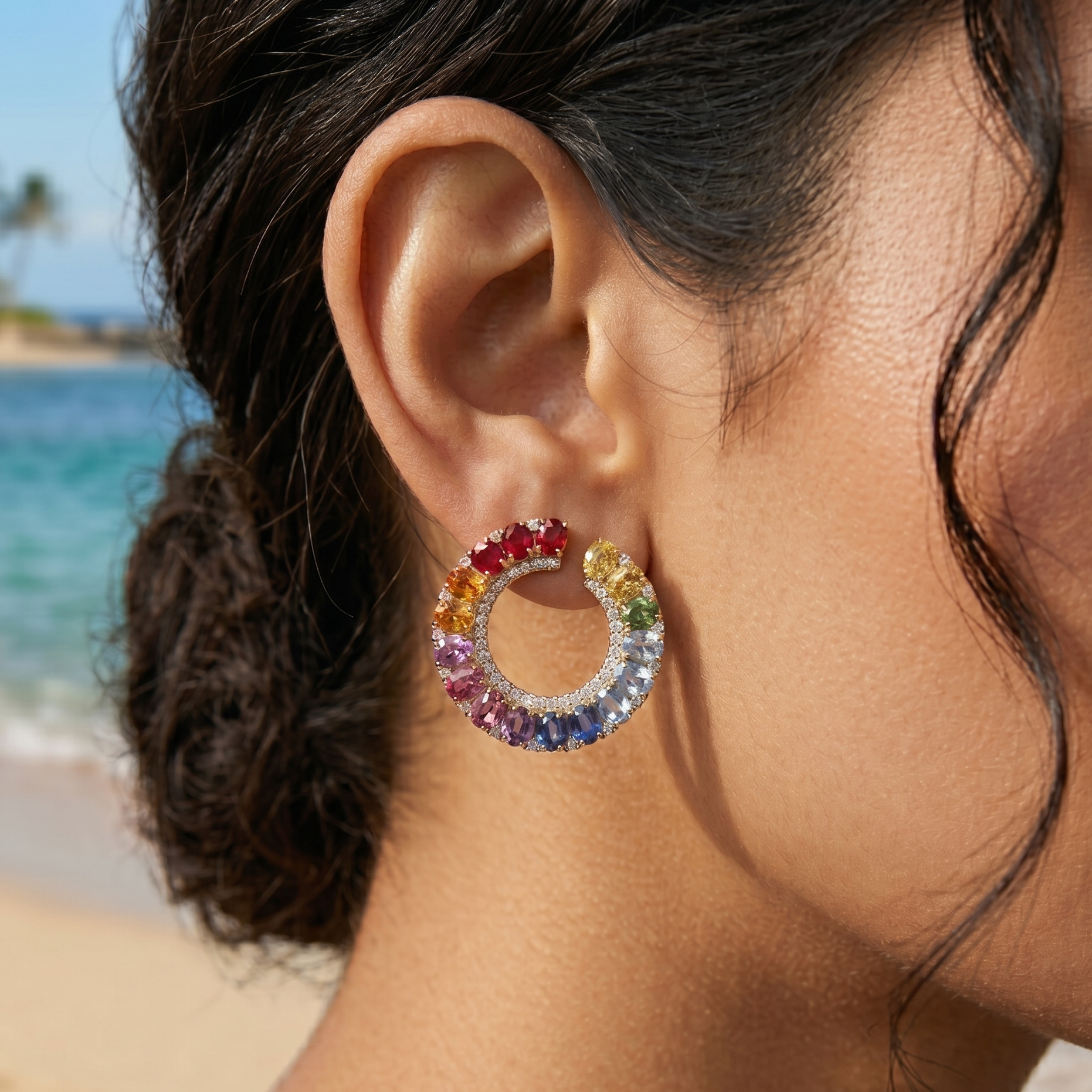 Close-up of a woman's ear wearing a colorful rainbow/multi sapphire diamond hoop earring with a beach in the background 14 kt gold delhi the alchemy studio