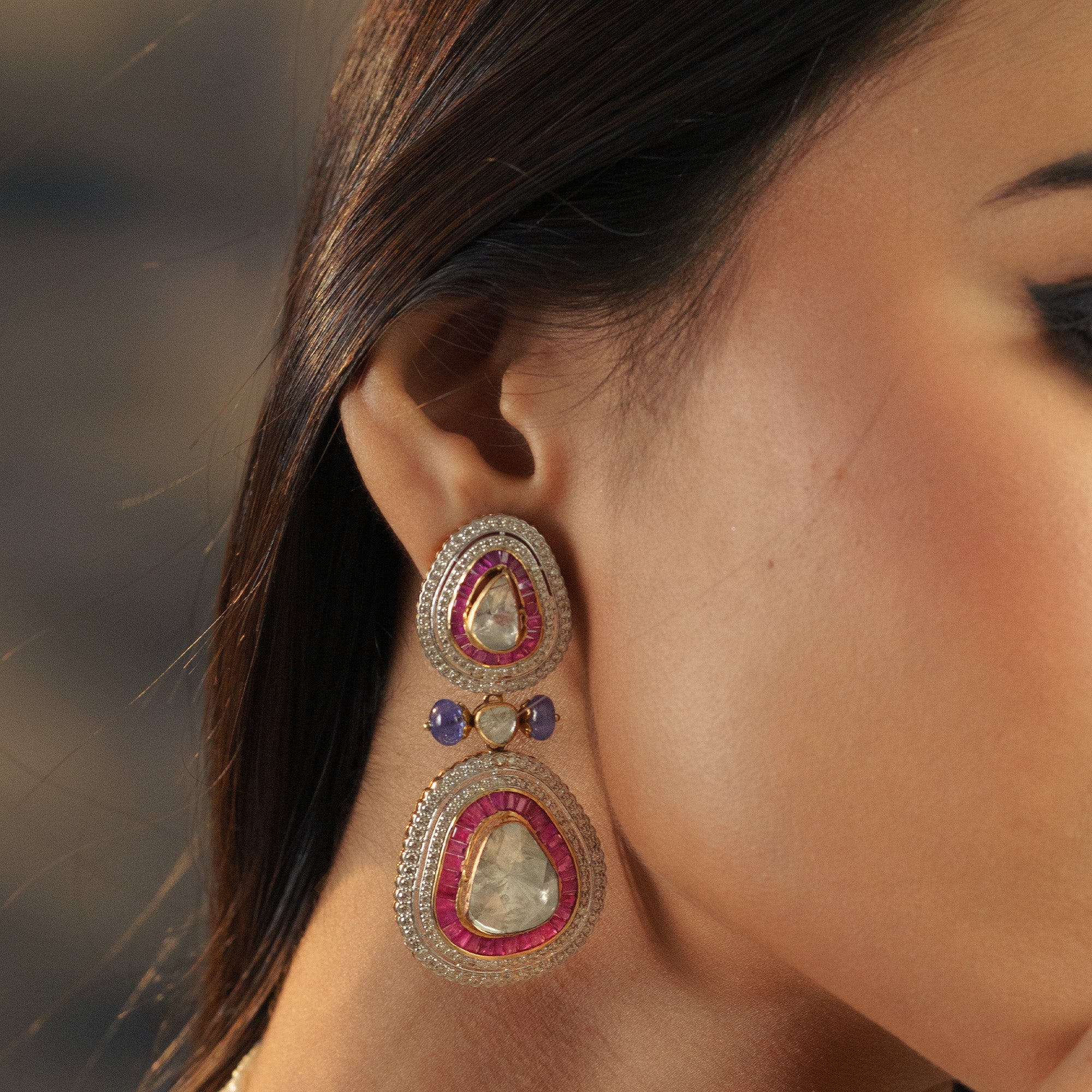 Side profile of a model wearing gold Polki diamond drop earrings featuring pear-shaped uncut diamonds surrounded by a baguette-set ruby halo and sapphire accents in india