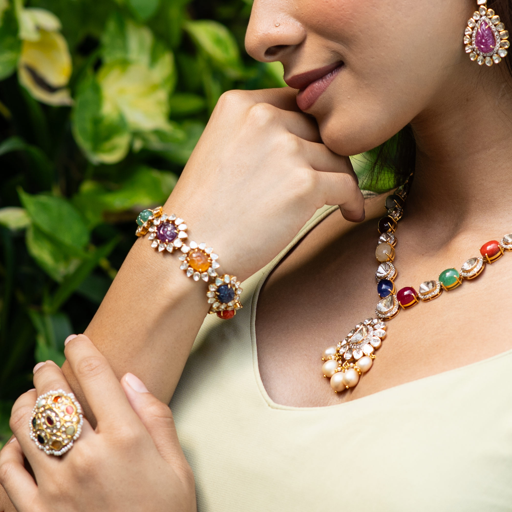 A close-up lifestyle shot of a woman wearing an ornate gold jewelry set with multicolored gemstones, including a ring, bracelet, necklace, and matching earring, set against a soft-focus background of green leaves.
