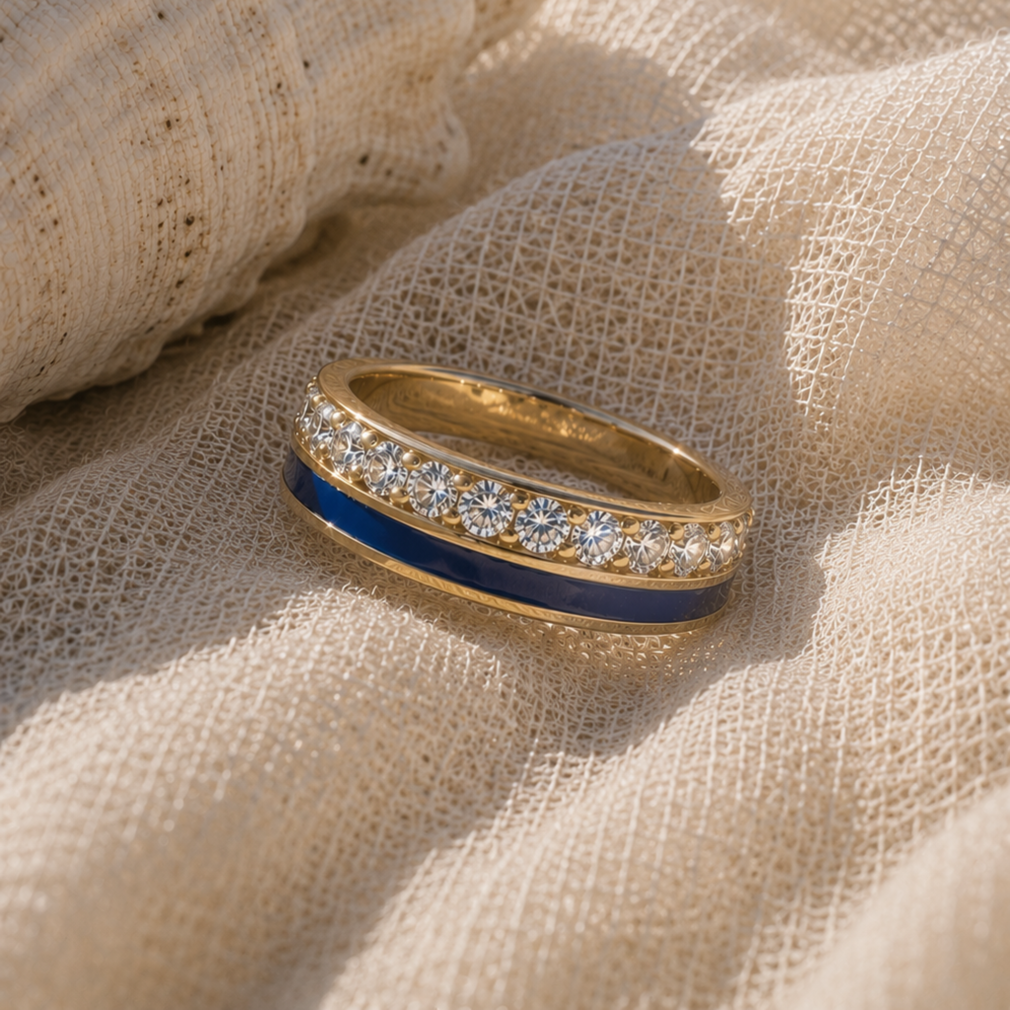 A gold band ring featuring a deep blue inlay and a row of clear pavé-set round stones, resting on a textured beige linen fabric.