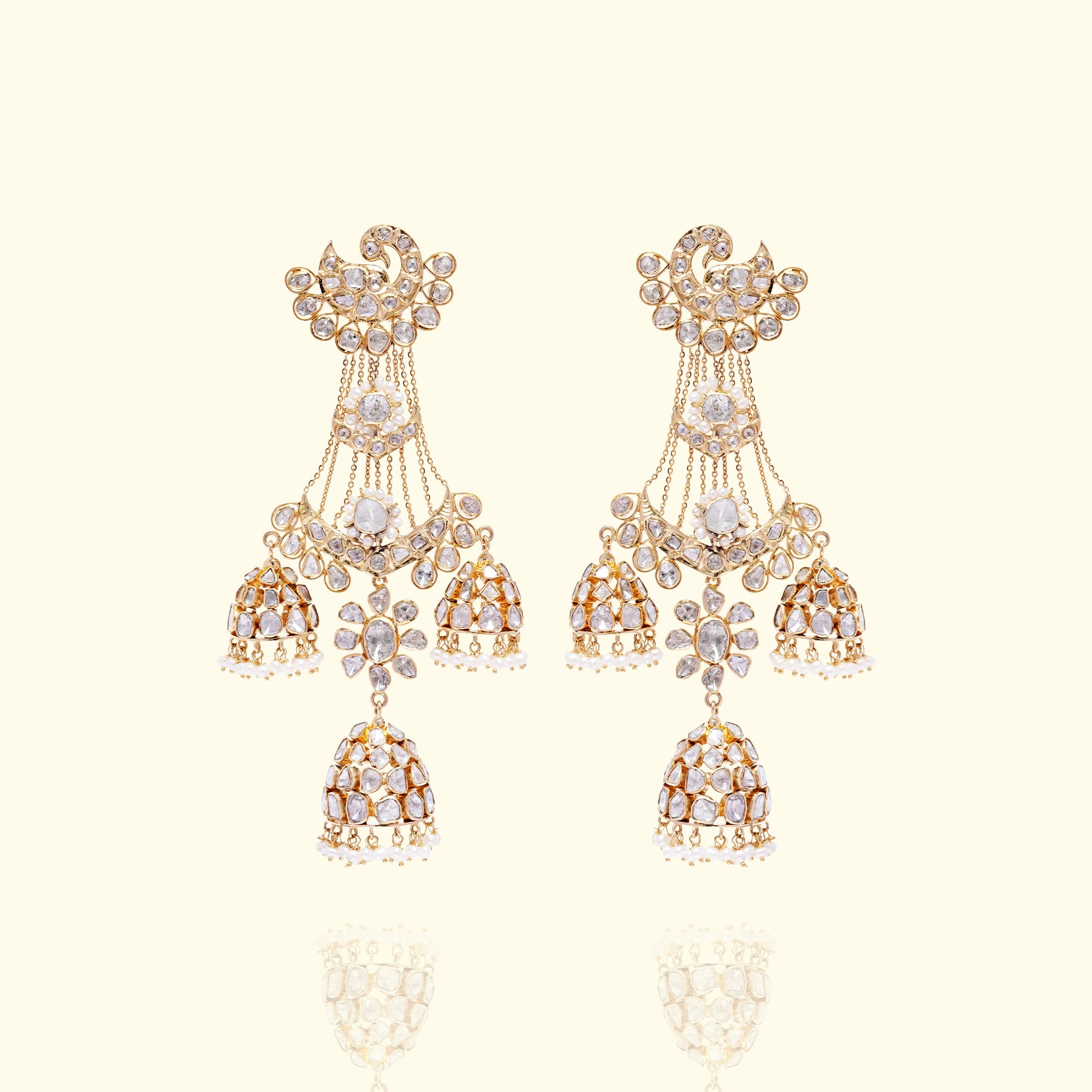 A pair of grand gold polki earrings featuring a peacock-shaped stud with uncut diamond plumage, supporting three tiered, bell-shaped jhumka drops suspended by delicate gold chains and finished with tiny japanese freshwater pearl fringes.