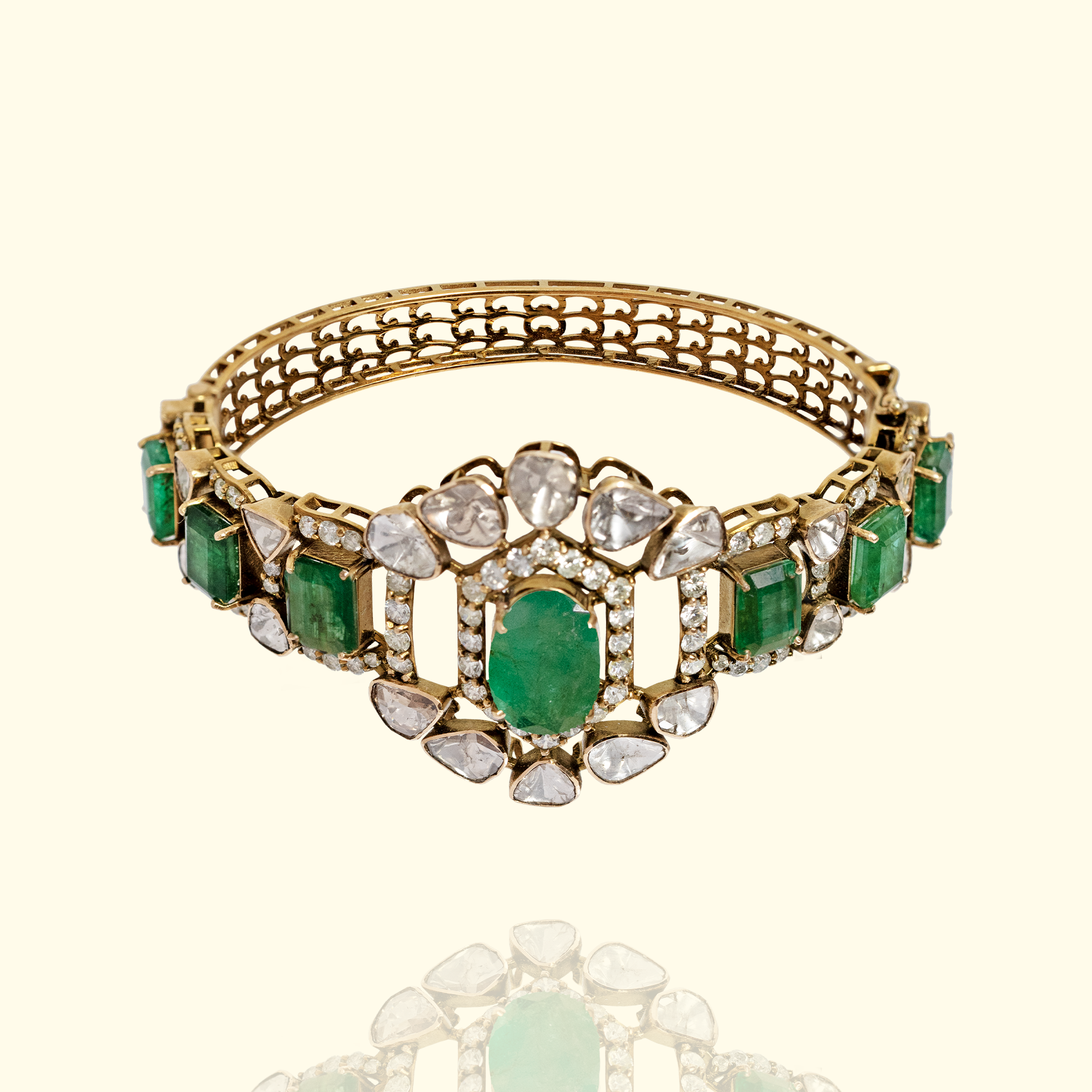 Studio shot of a high-jewelry gold Polki and emerald bangle with a central oval emerald, pear-shaped diamond accents, and an openwork filigree band.