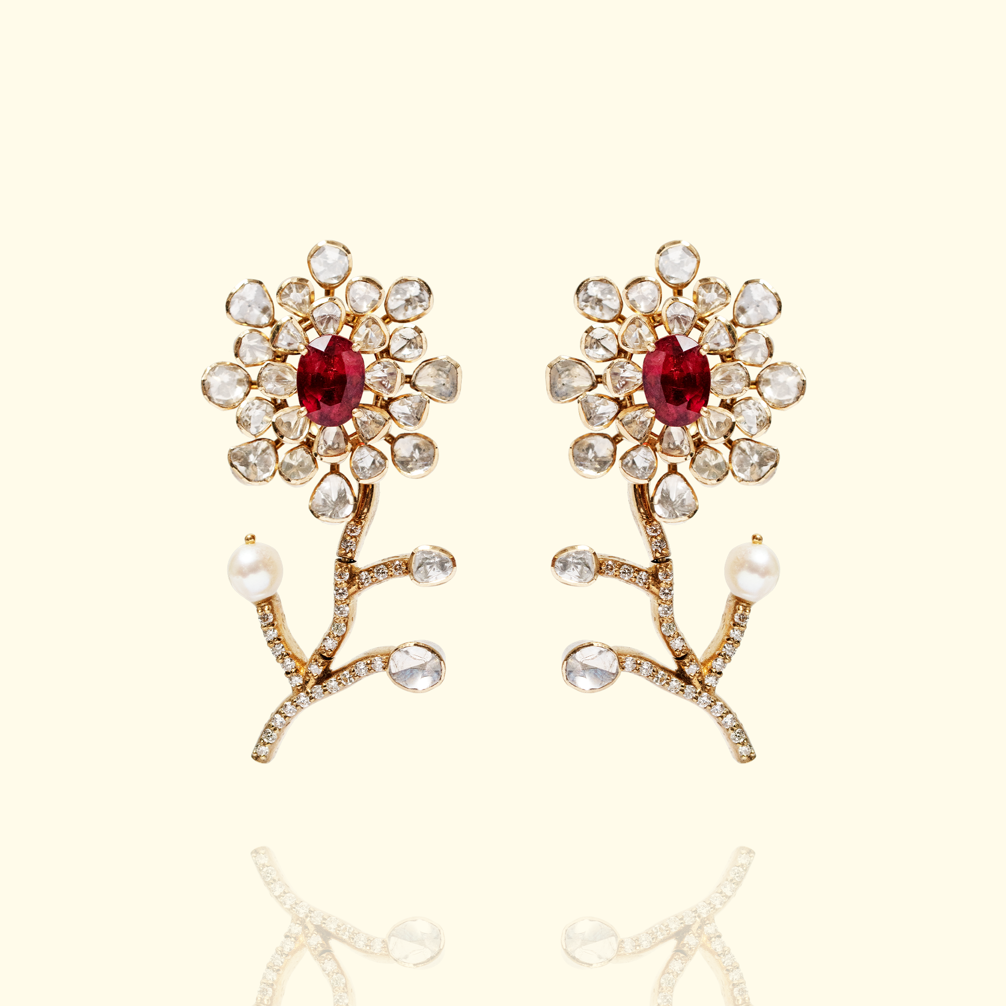 A pair of artistic gold polki ruby earrings shaped like delicate blooming flowers, featuring a central oval-cut ruby surrounded by a petal-like halo of uncut diamonds, set atop a curved stem accented with pavé diamonds and a single seed pearl.