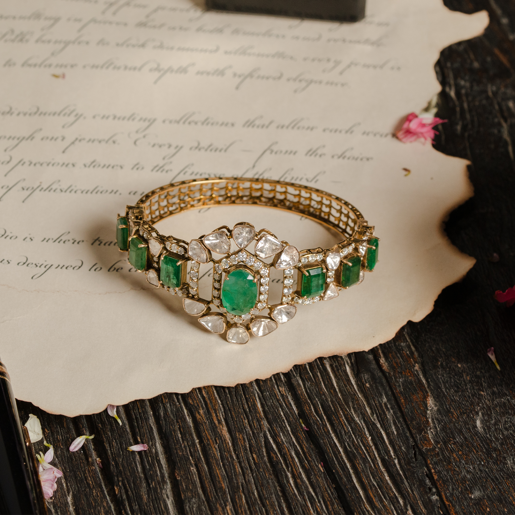 Ornate gold Polki and emerald statement bangle, bracelet featuring a central floral polki diamond emerald motif and a band of rectangular emeralds, displayed on vintage parchment.