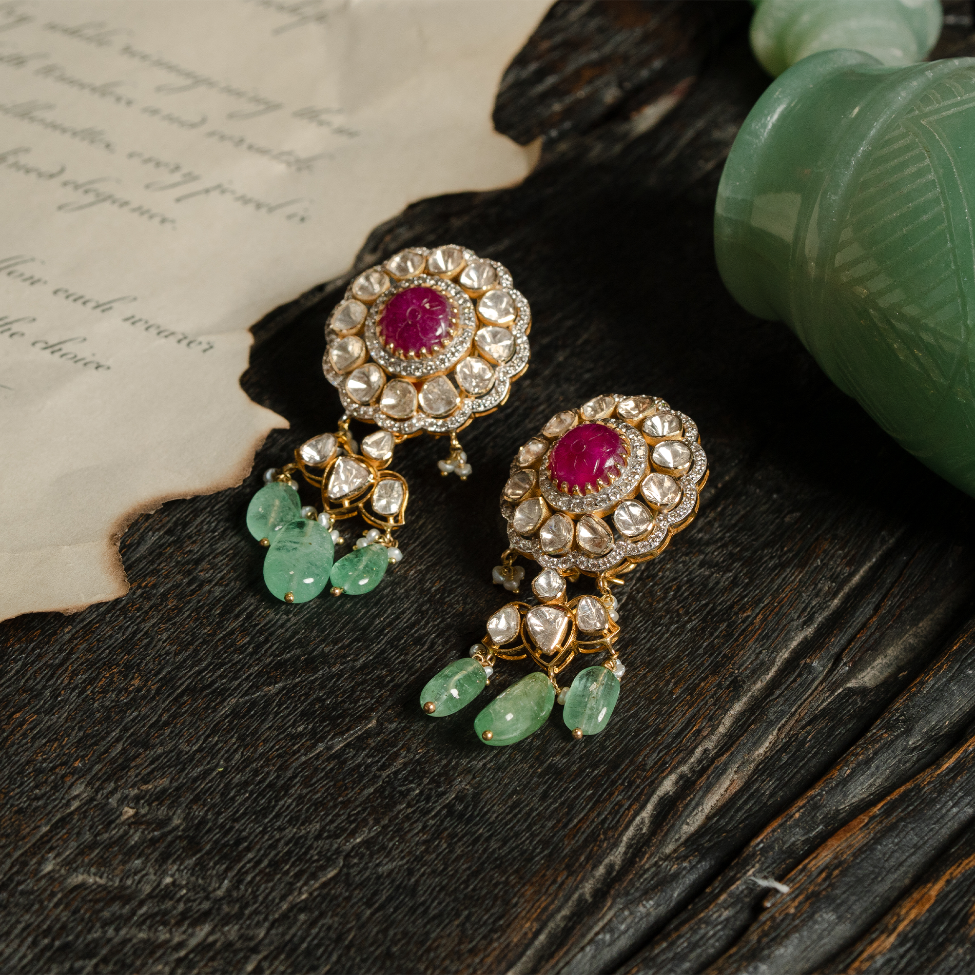 A pair of ornate floral-motif gold polki earrings featuring a central carved ruby cabochon surrounded by uncut diamonds and finished with cascading emerald droplets.