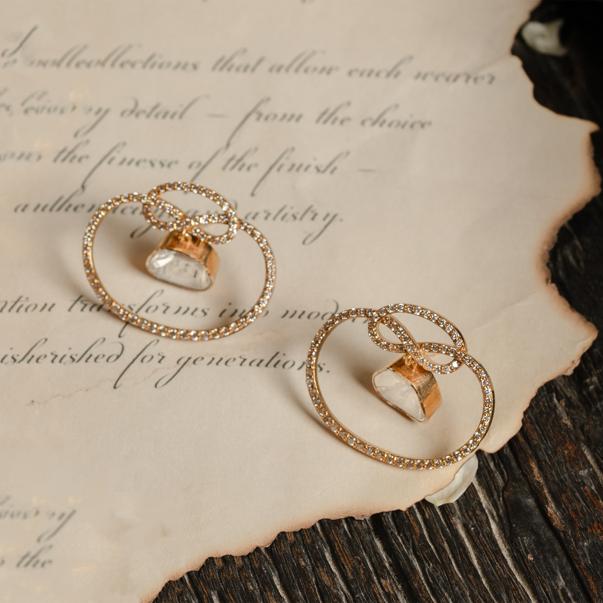 A pair of modern minimalist stud earrings featuring a central triangular polki diamond suspended within a delicate, round cut diamond-encrusted gold infinity knot.