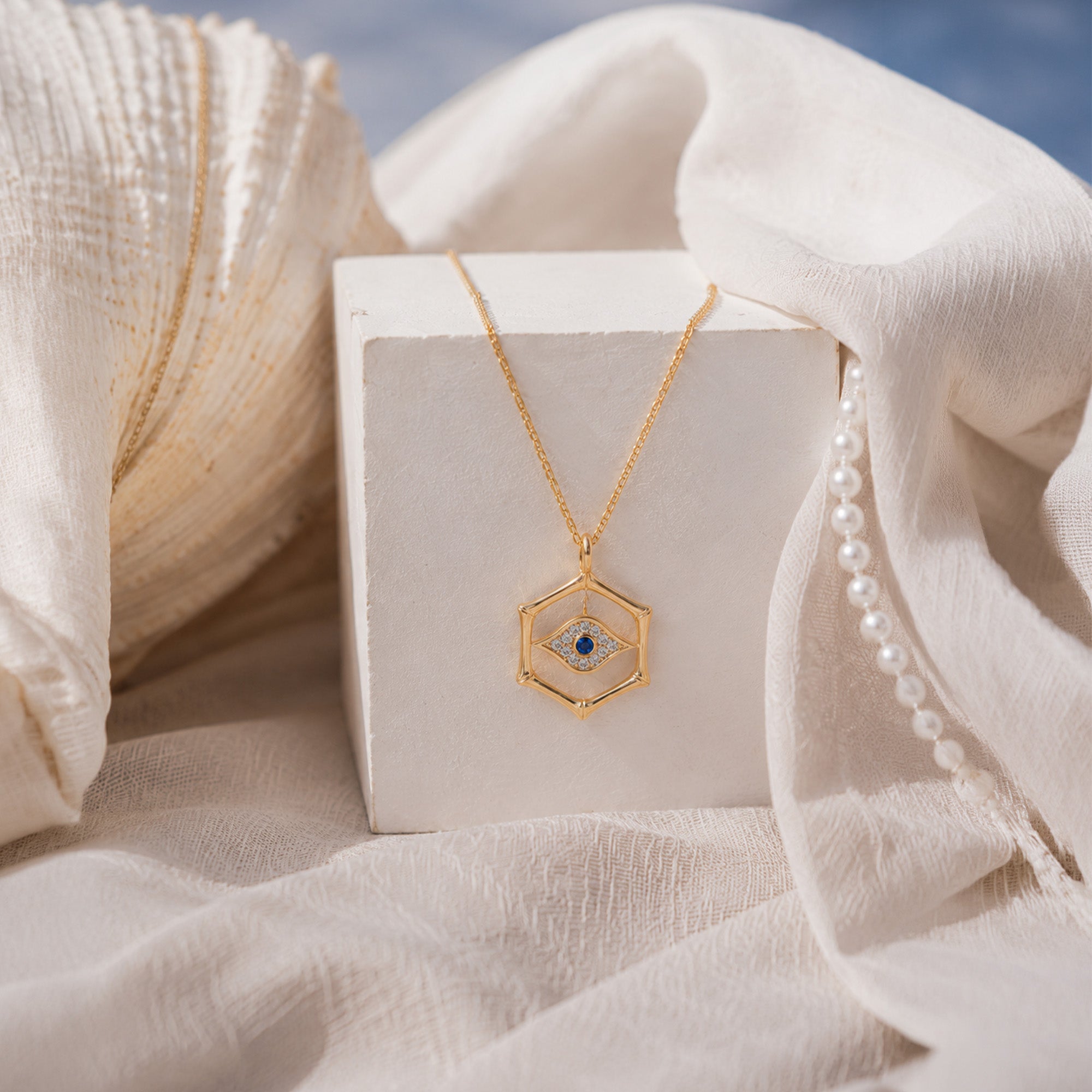 A gold hexagonal pendant necklace with a blue and white evil eye design, displayed on a white block surrounded by draped cream fabric and a pearl strand.