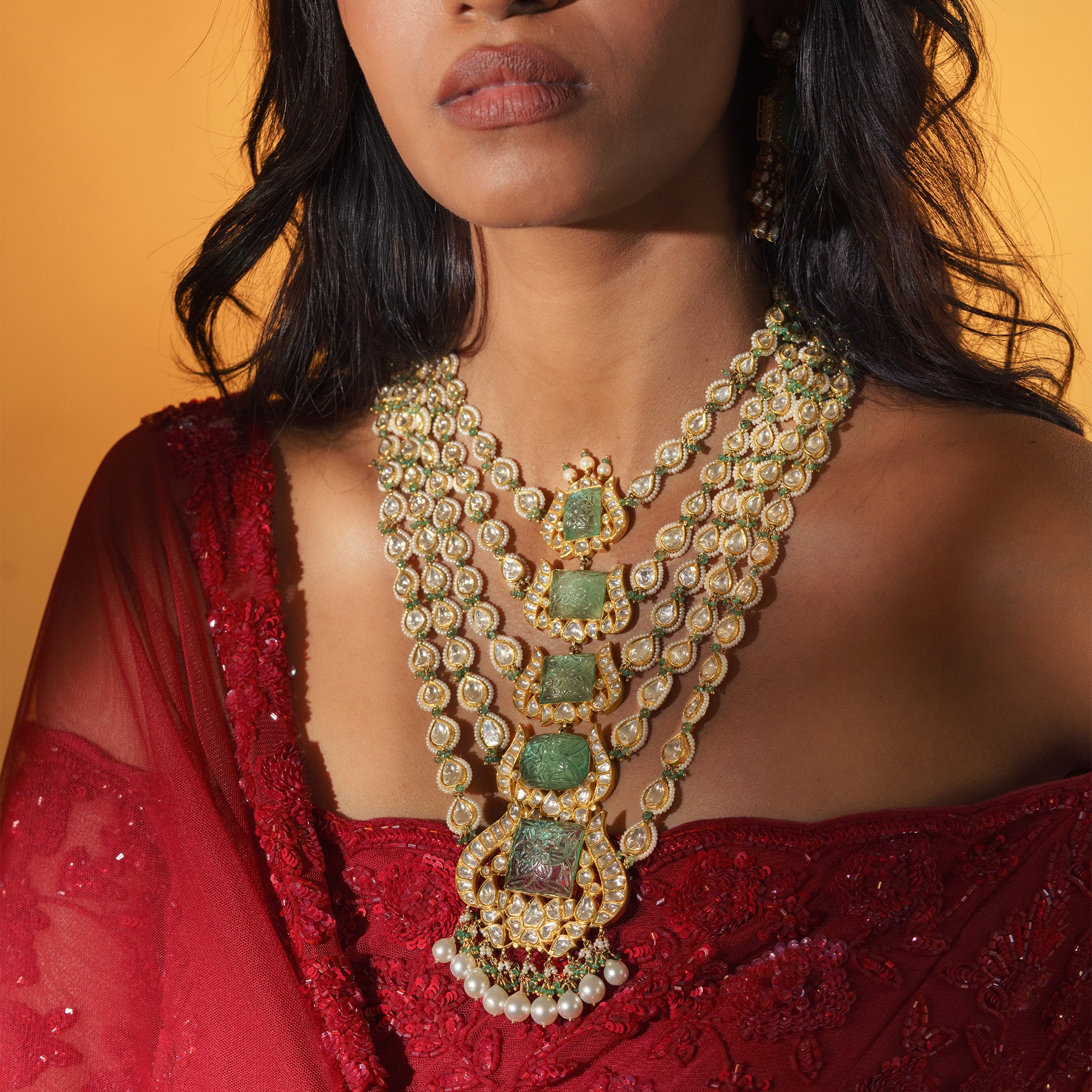 A model in red traditional attire wearing a magnificent five-strand Satlada necklace featuring graduated rows of teardrop polki diamonds and a vertical line of five carved emerald plaques in ornate gold settings.