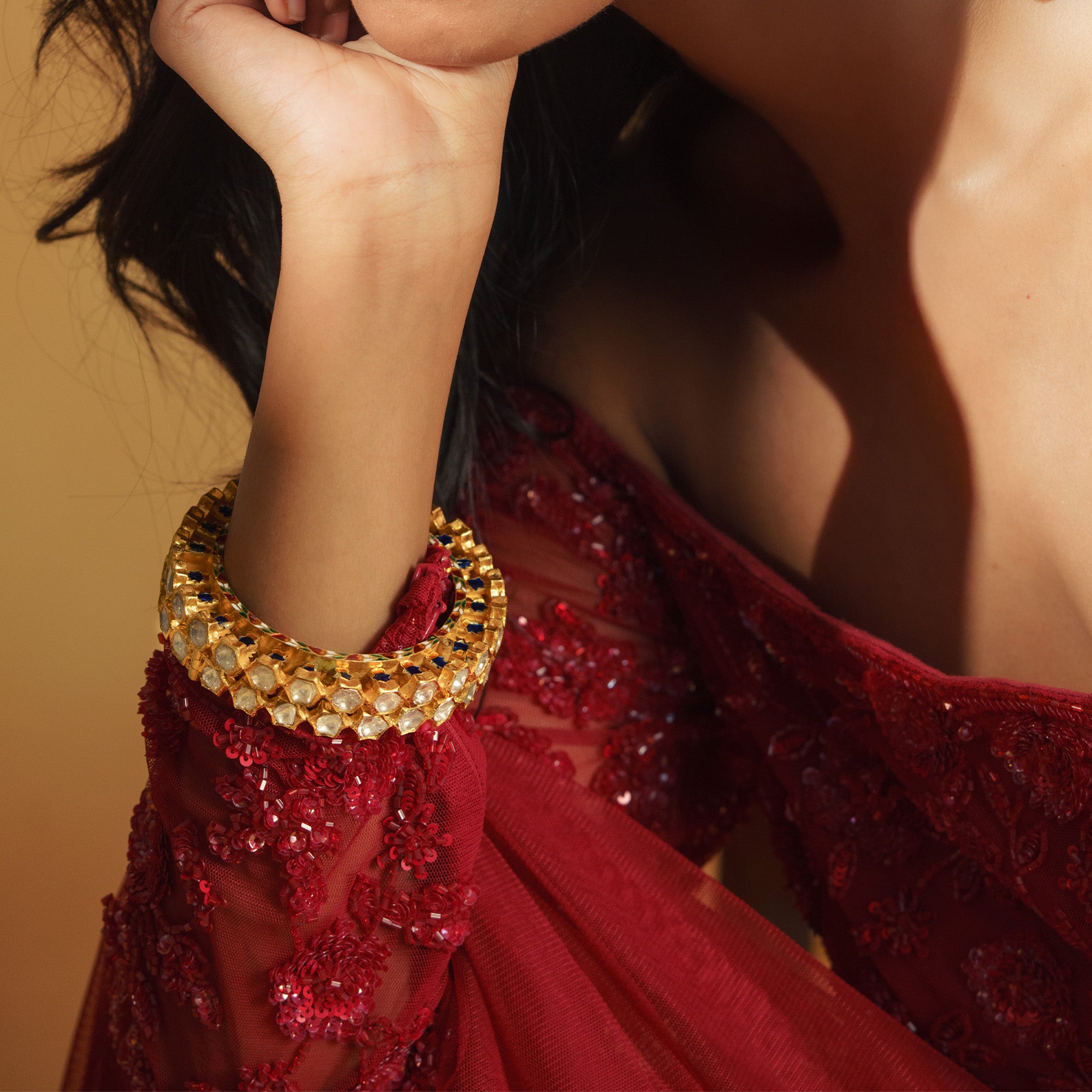 Close-up of a wide, ornate gold Polki diamond bangle with scalloped edges and green, blue enamel accents, styled over a red embroidered sleeve.