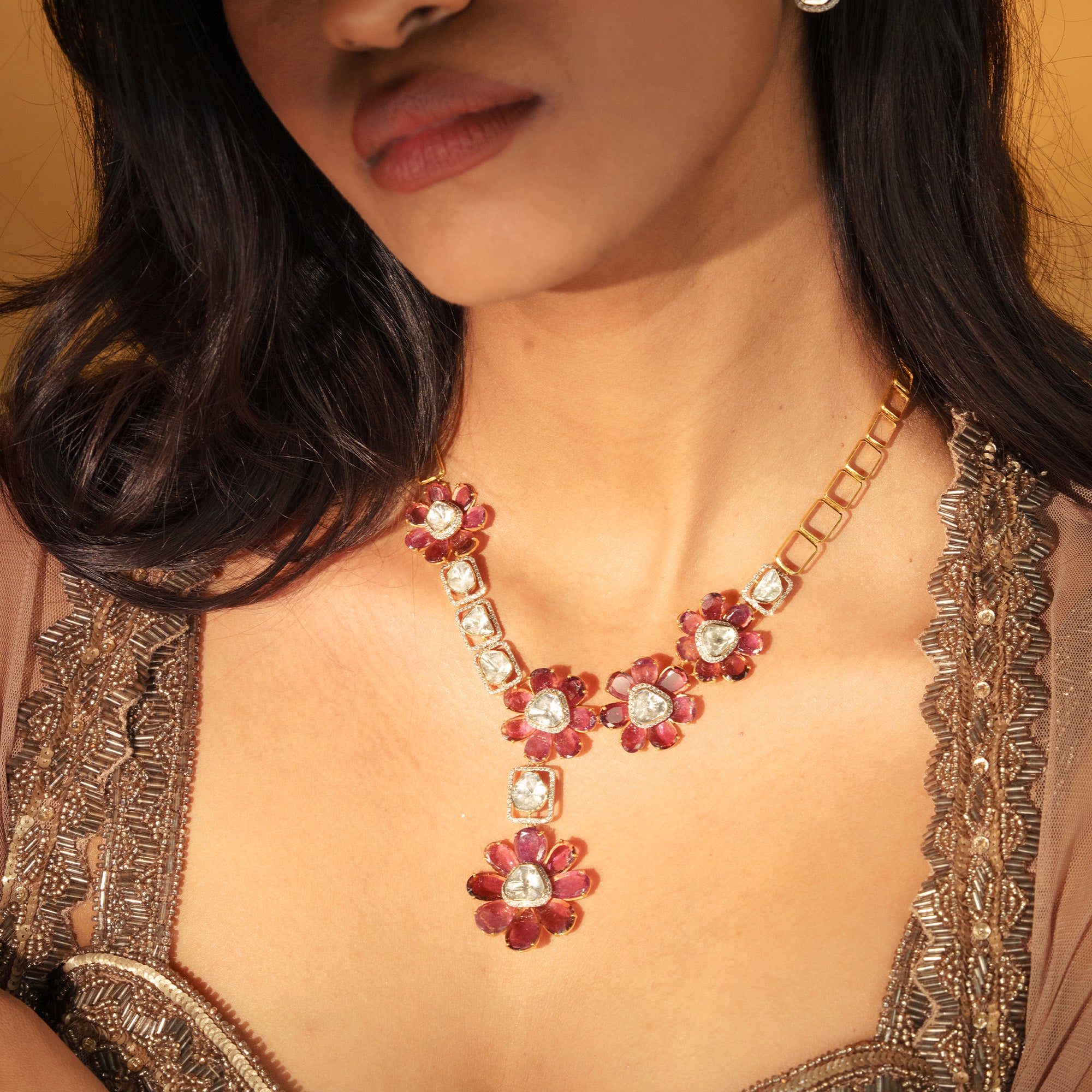 A model wearing a contemporary gold polki necklace featuring an asymmetrical arrangement of floral motifs with vibrant tourmaline petal and central polki diamonds, interspersed with geometric rectangular links encrusted with pavé diamonds and additional uncut diamonds.