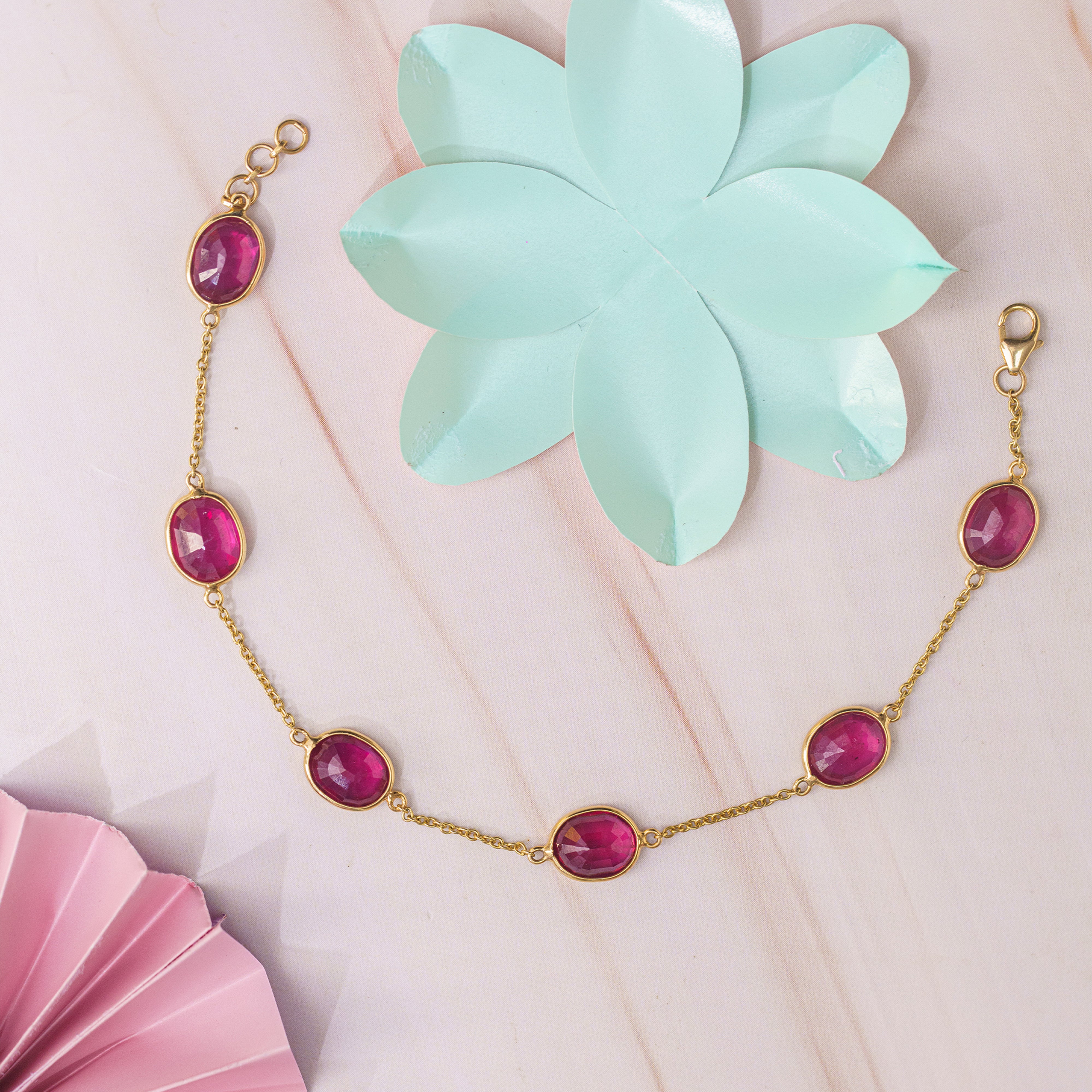 Gold chain bracelet with five oval pink ruby gemstones, styled on a light pink background with a mint green paper flower and a pink folded fan.