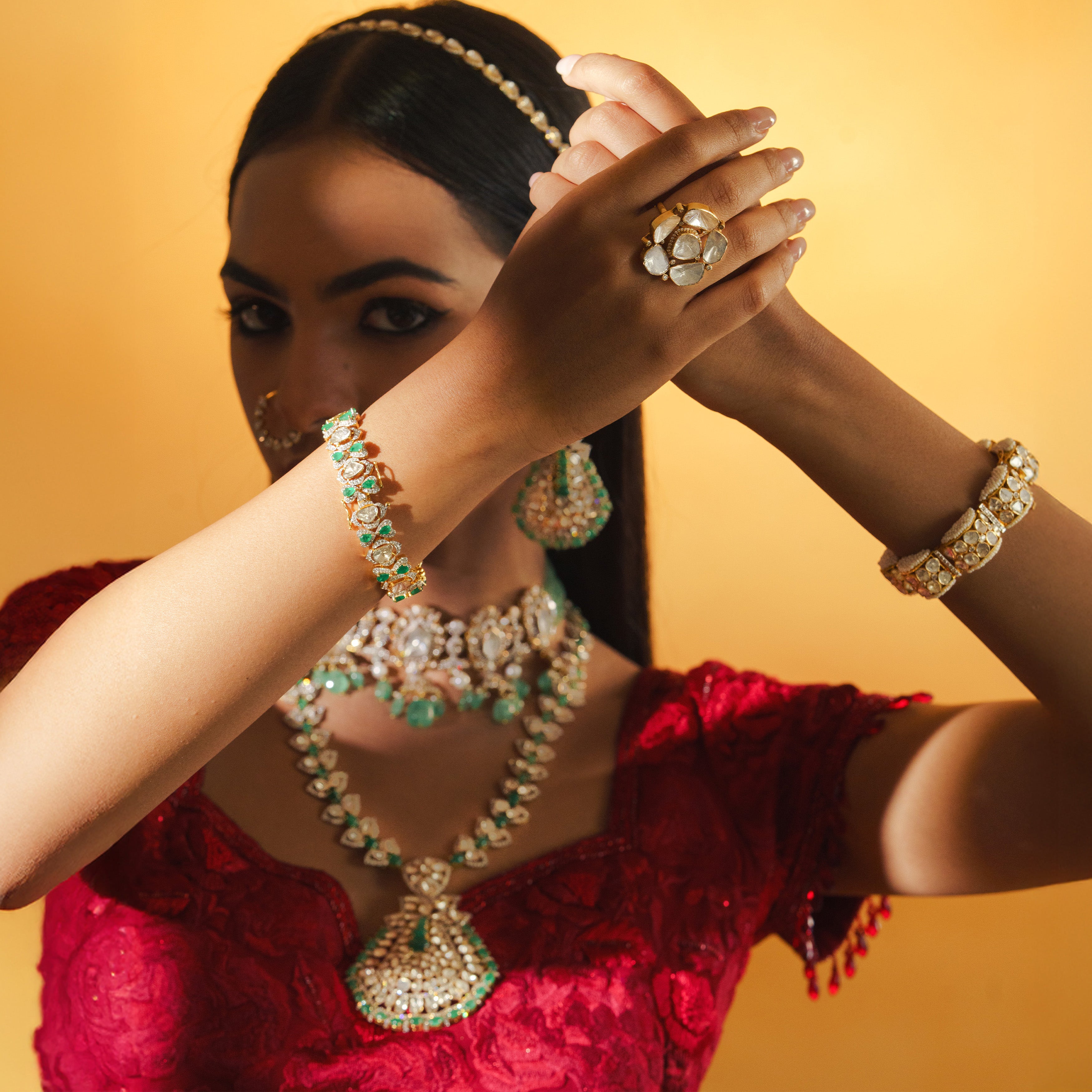 A model in a red ensemble showcasing a layered jewelry set, including a mint-green emerald bead choker and a long polki diamond necklace with a large fan-shaped pendant, complemented by a floral polki statement ring and coordinating uncut diamond and emerald style bracelets.