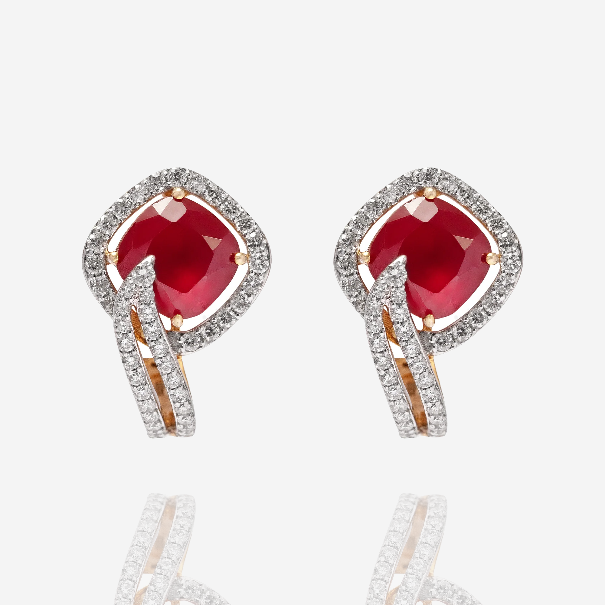 A pair of elegant ruby stud earrings featuring large cushion-cut red rubies set in gold, surrounded by a halo of pave diamonds with a modern, sweeping diamond-encrusted bypass detail in india
