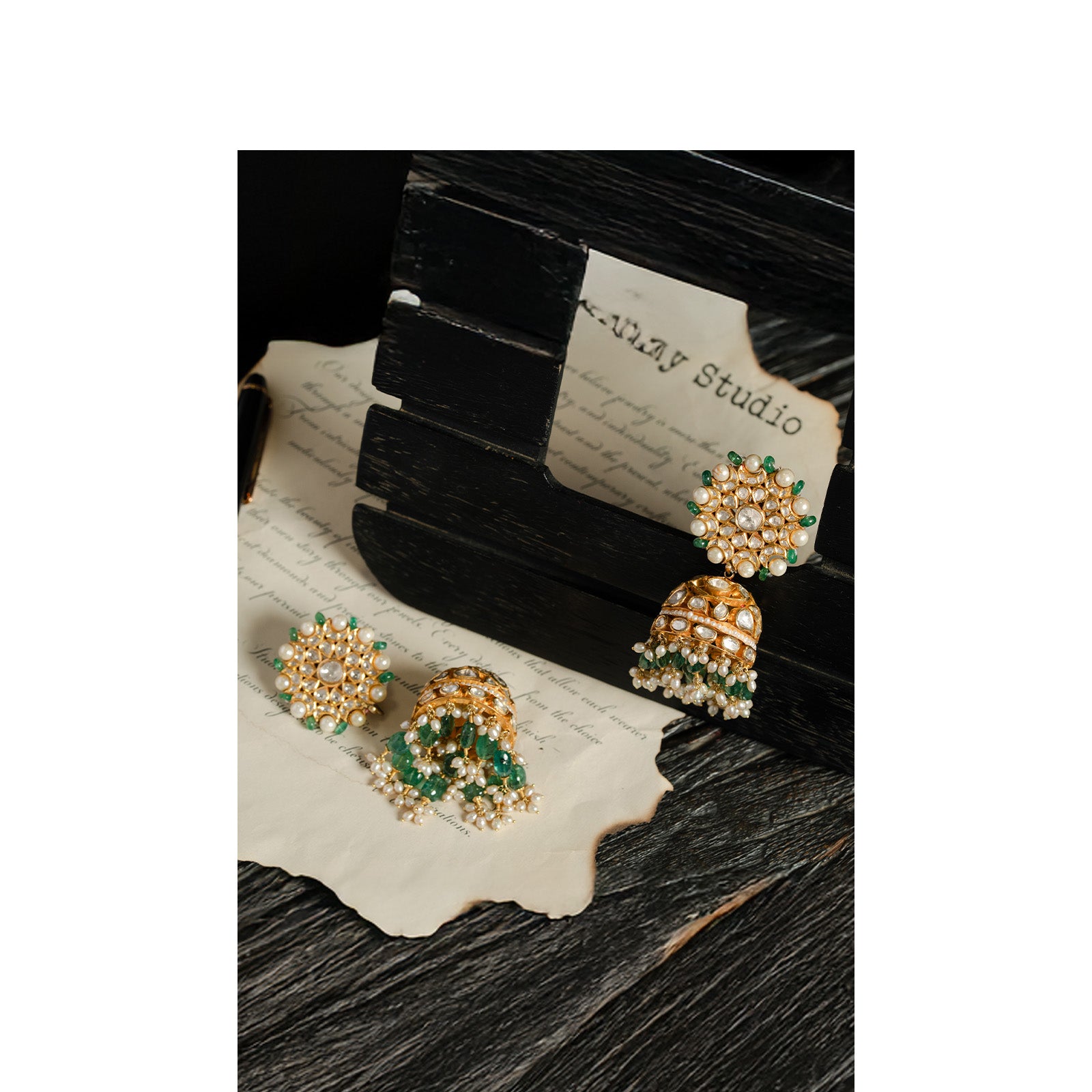 A pair of ornate gold jhumka earrings featuring large, circular floral studs encrusted with polki diamonds and a border of emerald beads, supporting bell-shaped drops adorned with rows of emeralds and delicate seed pearl fringes.