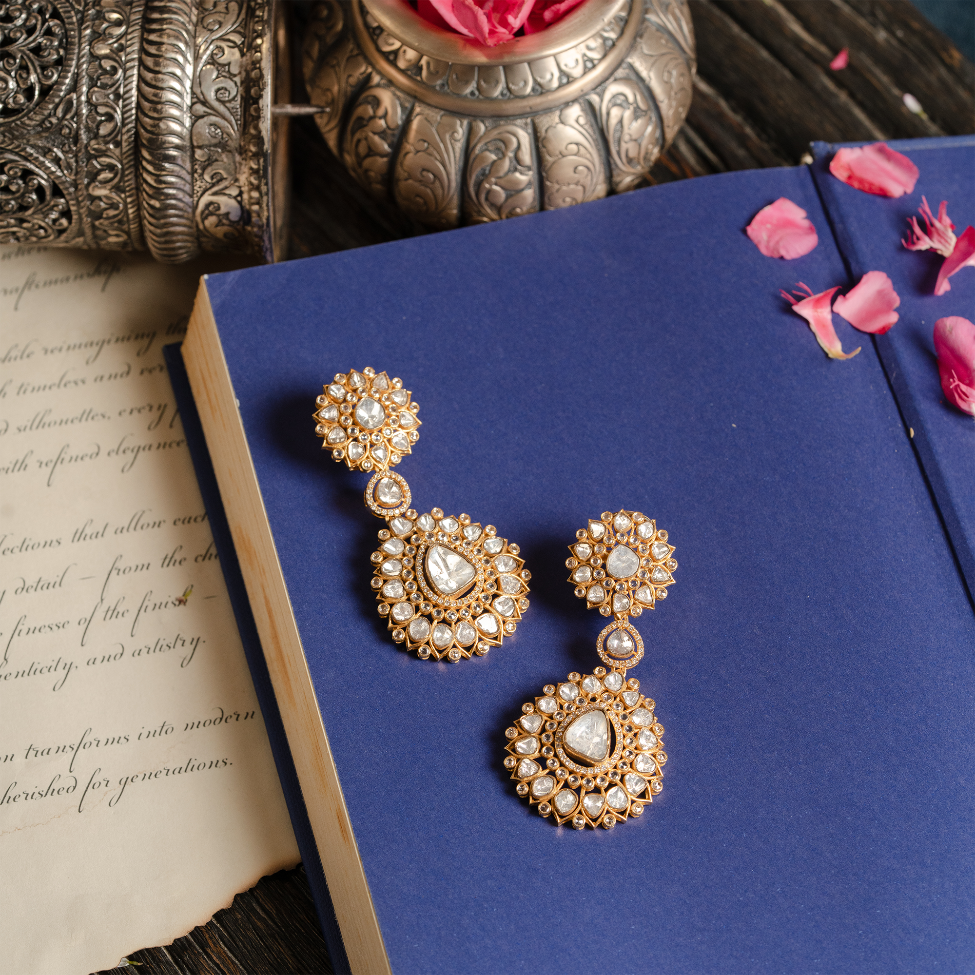 Pair of gold Polki diamond drop earrings featuring floral-motif studs and pear-shaped dangles, styled on a blue book with vintage parchment.