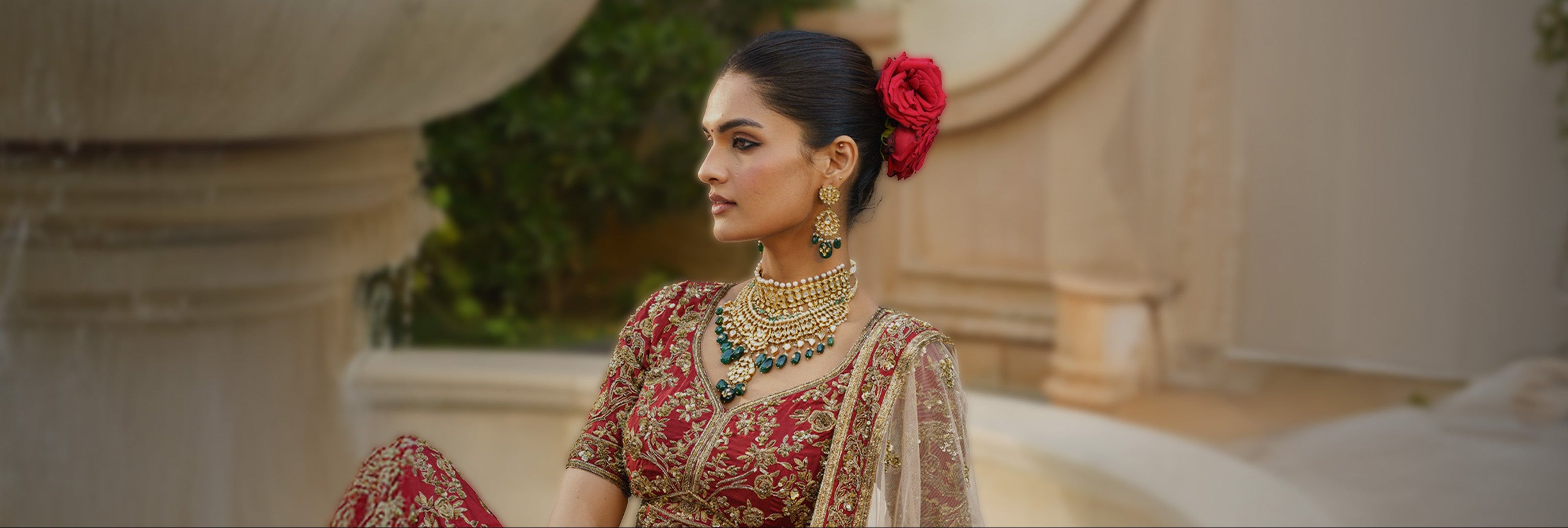 A model in a crimson embroidered lehenga wears a grand triangular polki diamond choker with a lush emerald bead fringe, layered with a pearl-strand necklace supporting a large, shield-shaped carved emerald pendant. The look is accessorized with coordinated emerald drop earrings and a hair adorned with fresh red roses.