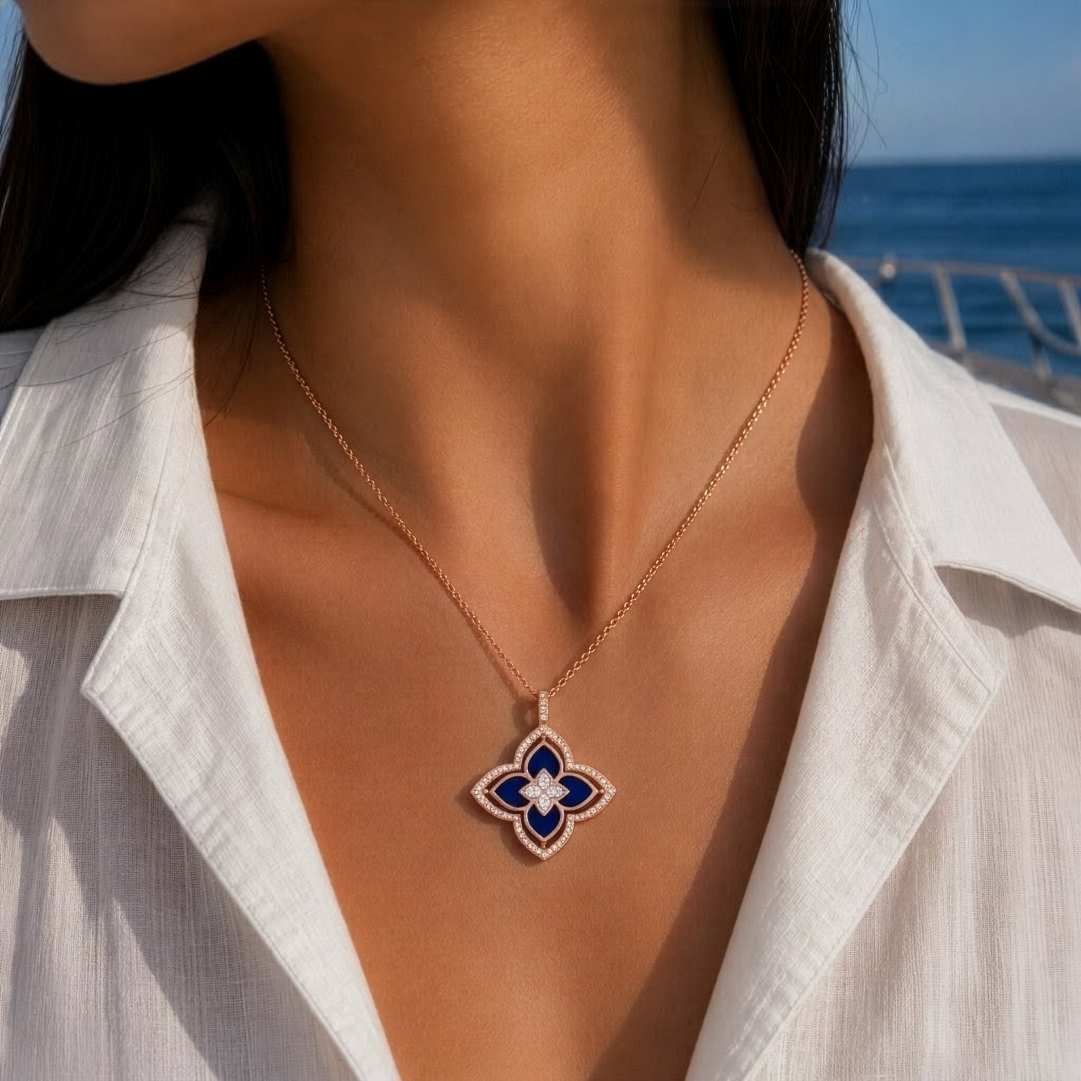 A close-up lifestyle shot of a woman wearing a delicate gold chain necklace with a four-petal clover pendant. The pendant features a blue center with a dark red outer border, worn over a white collared shirt with a blurred ocean background.