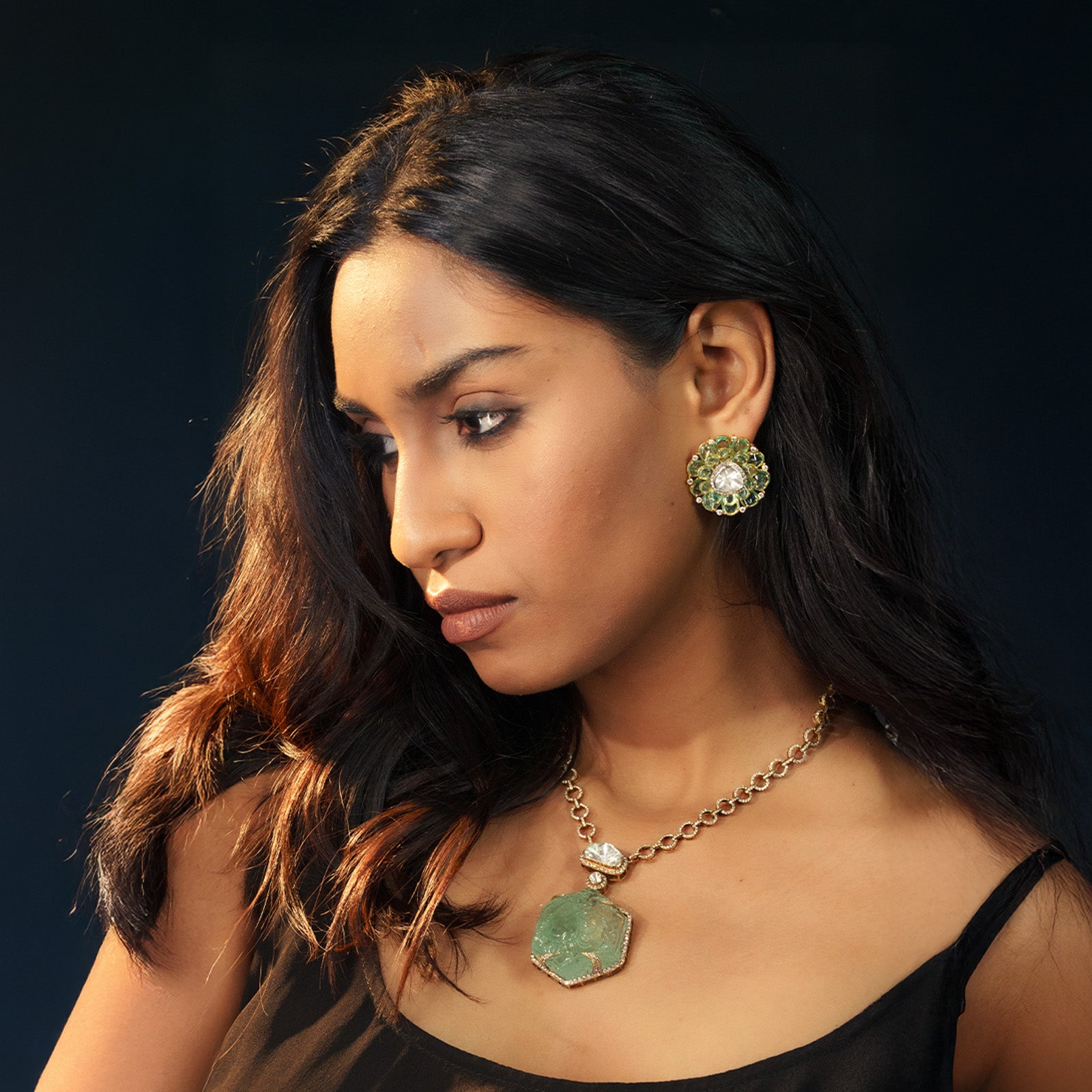 A model wearing a large, hexagonal carved emerald pendant suspended from a simple gold link chain, paired with matching circular emerald floral stud with central polki uncut diamond earrings.