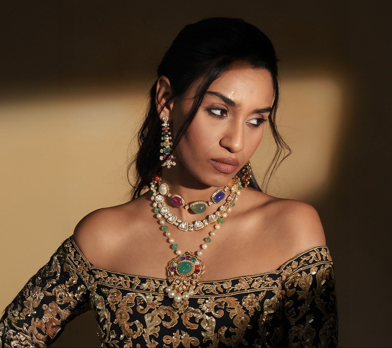 A model in a black and gold embroidered ensemble wearing a layered necklace set. The set includes a polki diamond choker accented with large emerald, ruby, and sapphire cabochons, and a long pearl strand necklace featuring a grand circular pendant with a carved emerald center, surrounded by a multi-colored navratan gemstone halo and pearl drops.