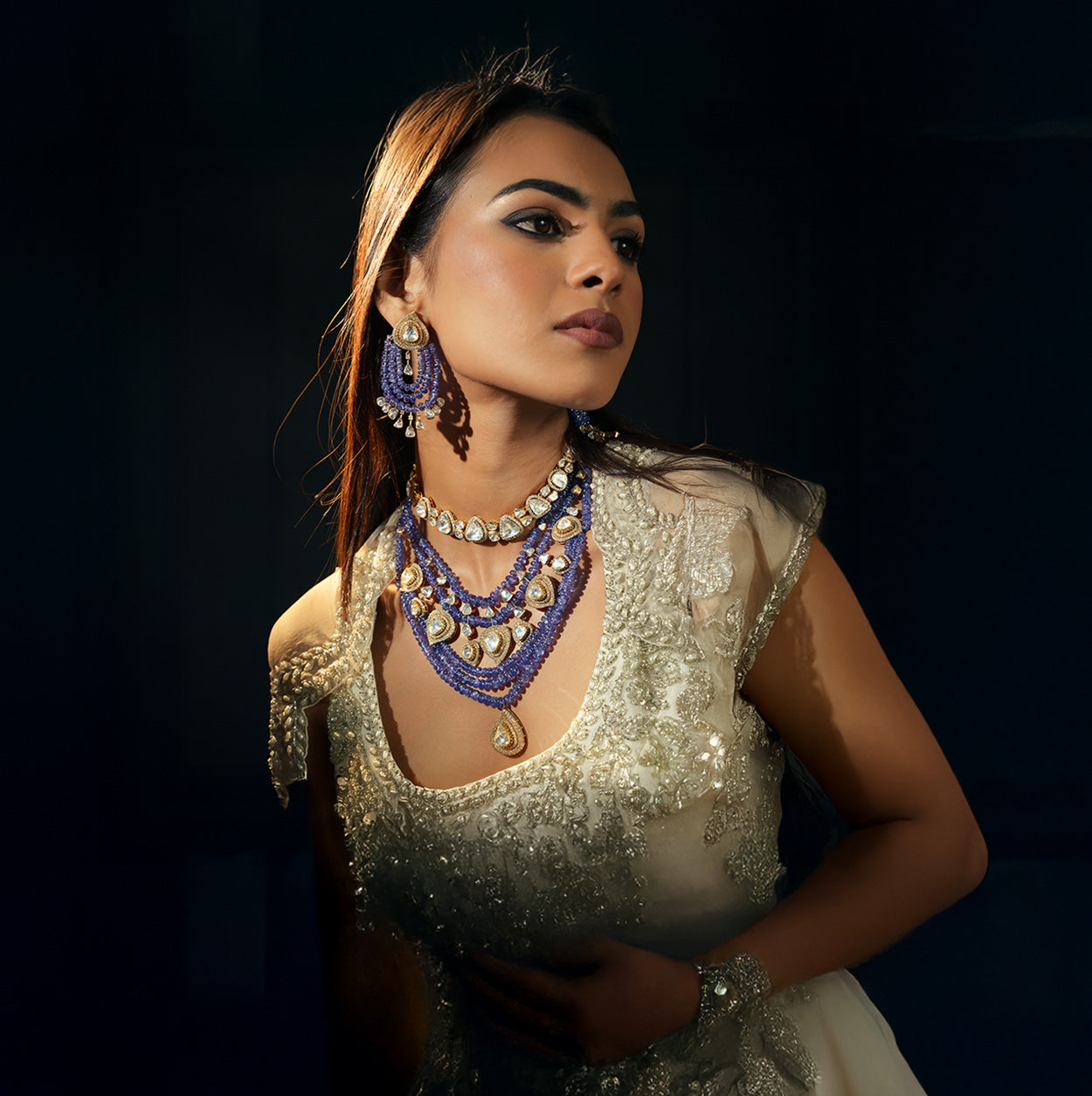 A model in a white embroidered gown wearing a layered polki diamond choker and a multi-strand blue tanzanite bead necklace featuring pear-shaped uncut diamond drops, paired with matching tanzanite bead chandelier earrings.