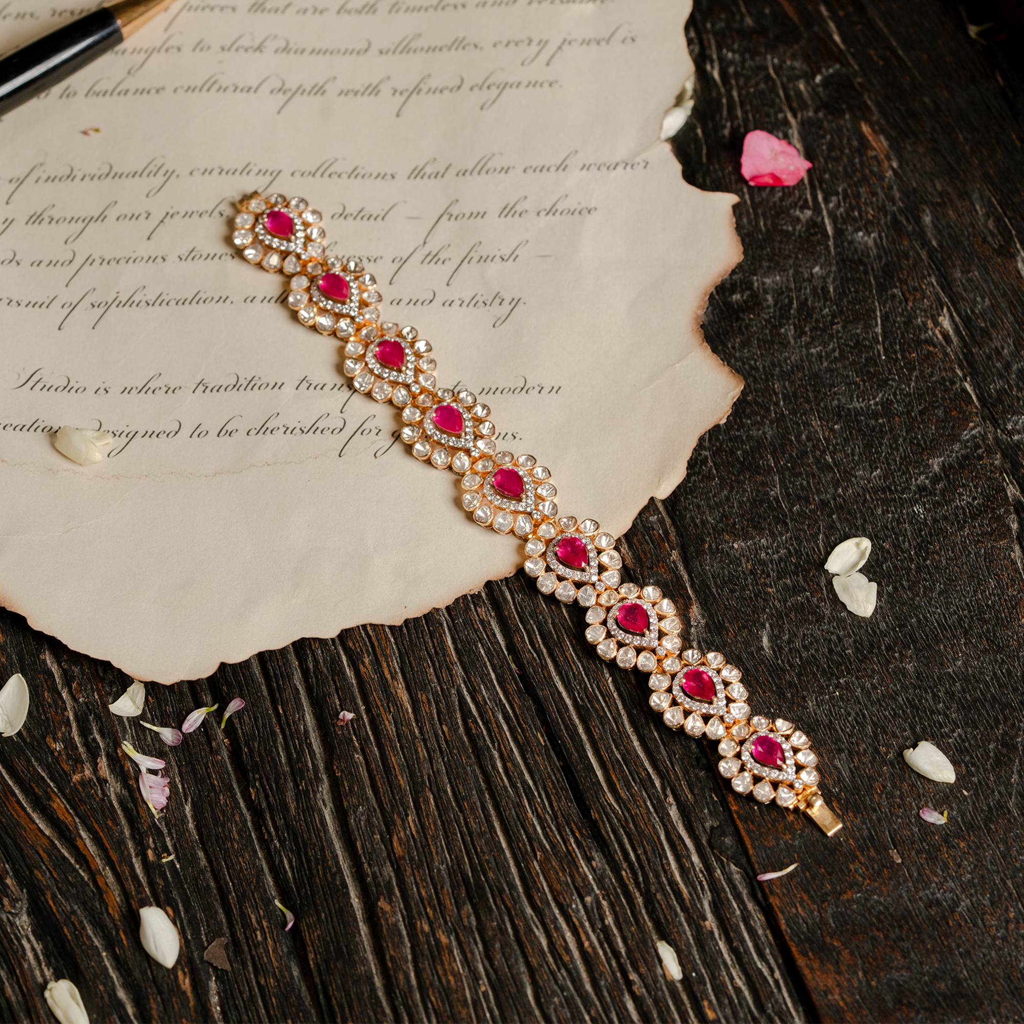Flat lay of a luxurious gold ruby and Polki diamond bracelet featuring pear-shaped rubies surrounded by uncut diamond halos, displayed on a piece of vintage parchment over a dark wooden surface with scattered flower petals.
