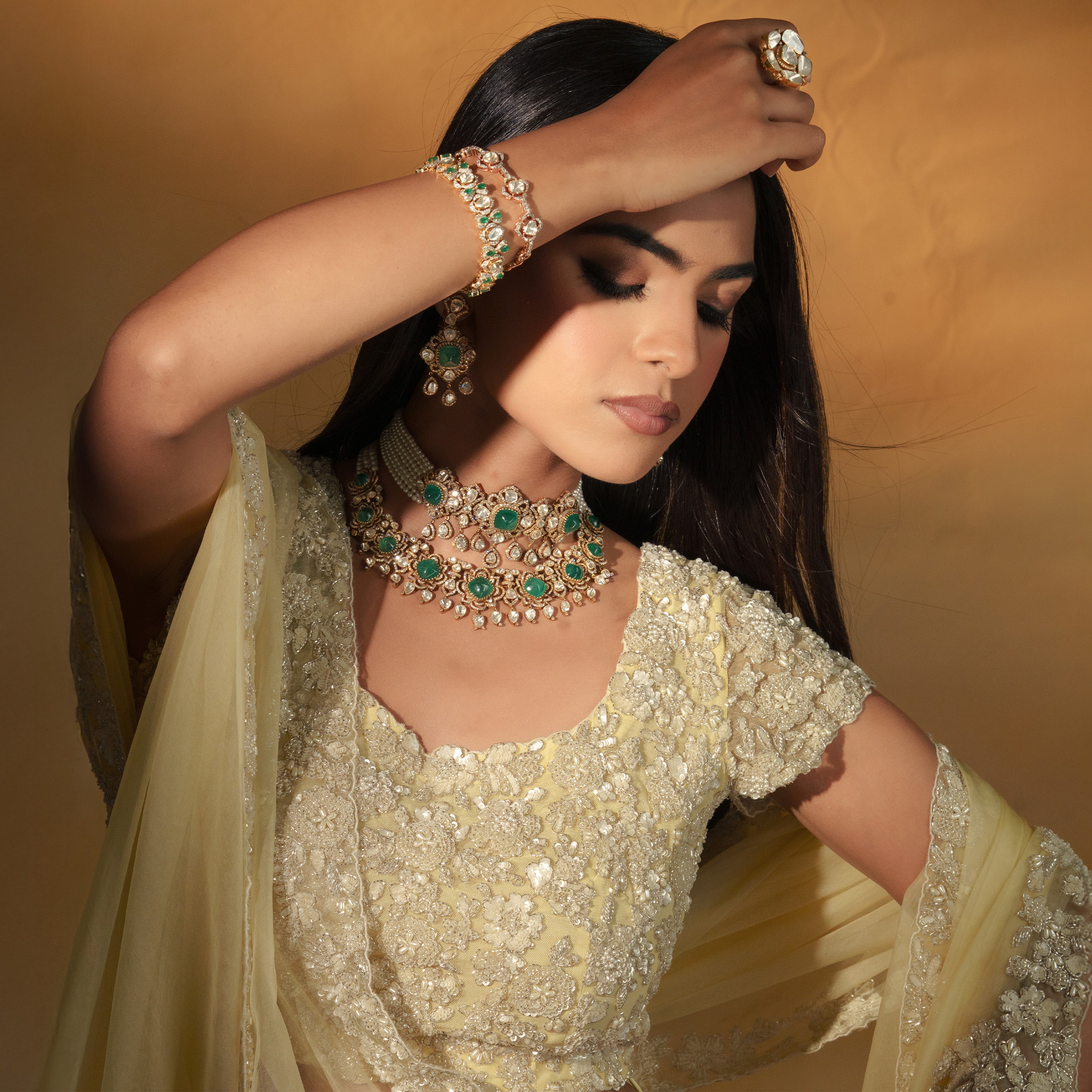Luxurious bridal jewelry set featuring a multi-layered Polki diamond choker with vibrant emerald centerstones, paired with a matching emerald bracelet and a statement Polki diamond ring, styled with a light yellow embroidered lehenga.