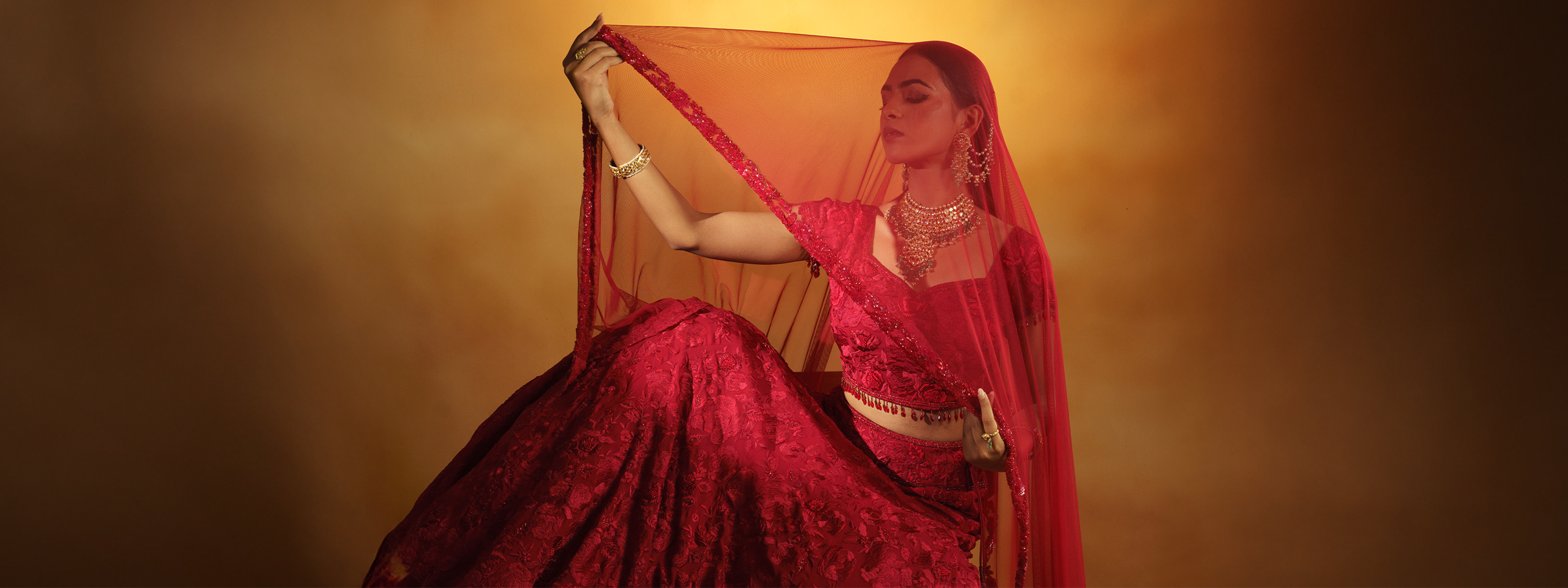 A model in a traditional red bridal lehenga wearing a heavy polki diamond choker with emerald drops, paired with matching layered sahara earrings and stacked gold bangles.