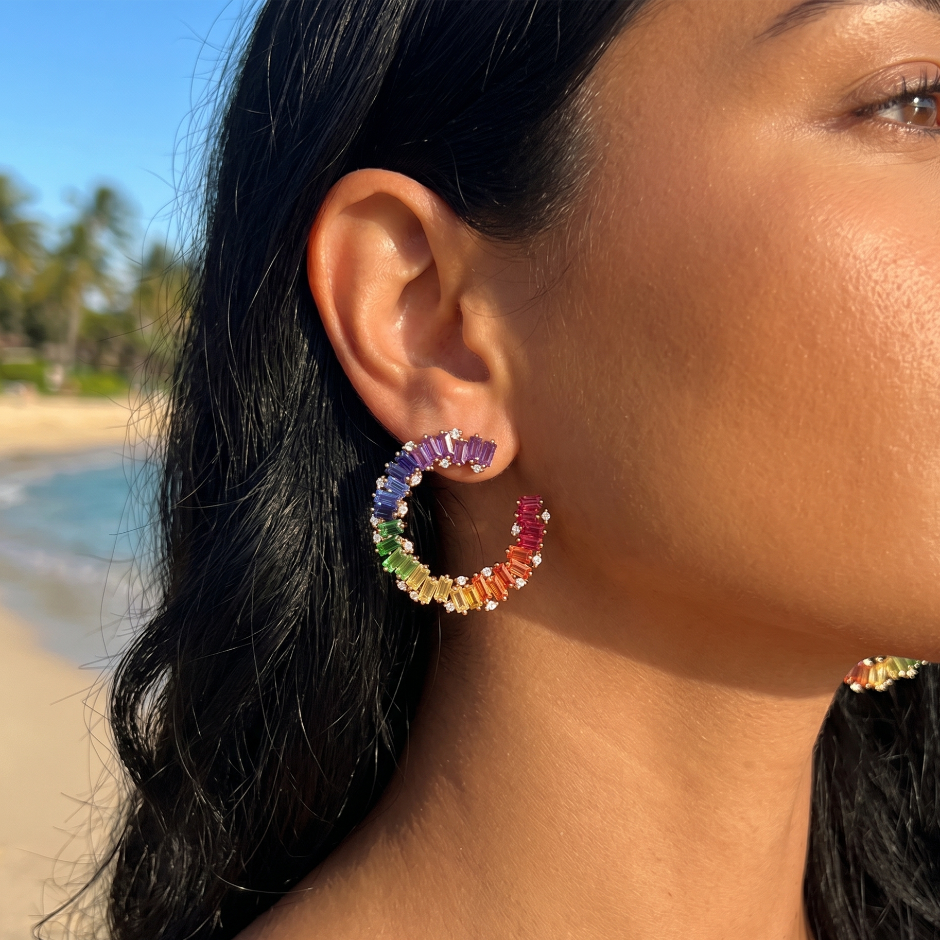 Close-up of a woman's ear wearing colorful baguette rainbow/multi sapphire diamond hoop earrings with a beach in the background 14kt white gold delhi the alchemy studio