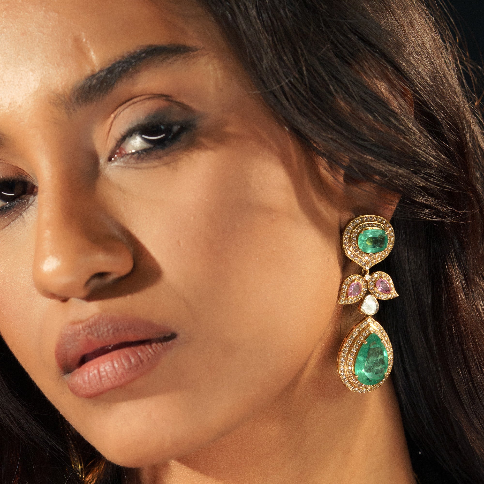 A model wearing regal triple-tier drop earrings featuring a round emerald stud, a leaf-motif center with tourmaline, and a large pear-cut emerald drop, all encased in shimmering brilliant cut diamond halos in india