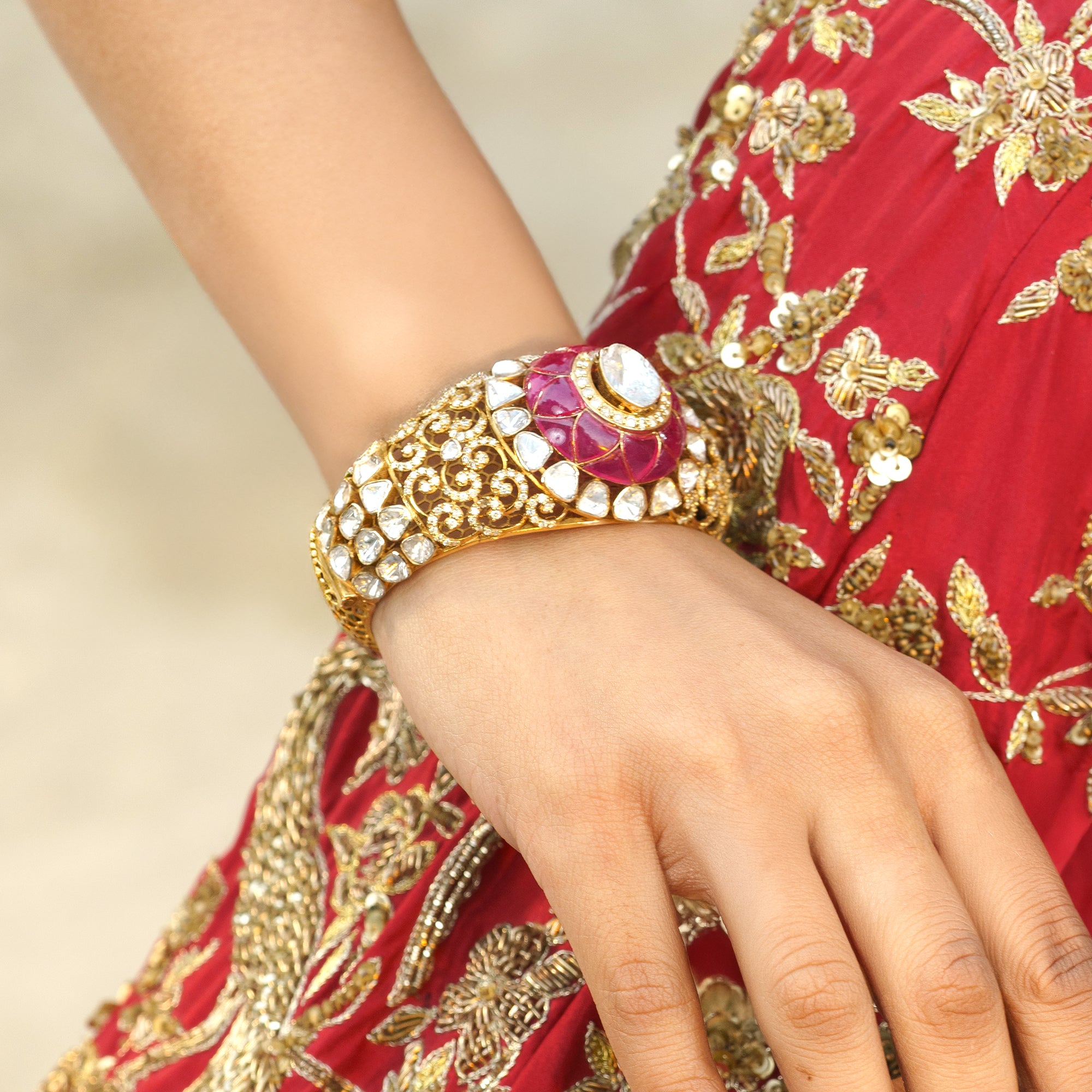 Gold bracelet with a red gemstone on a hand wearing a red and gold embroidered garment.