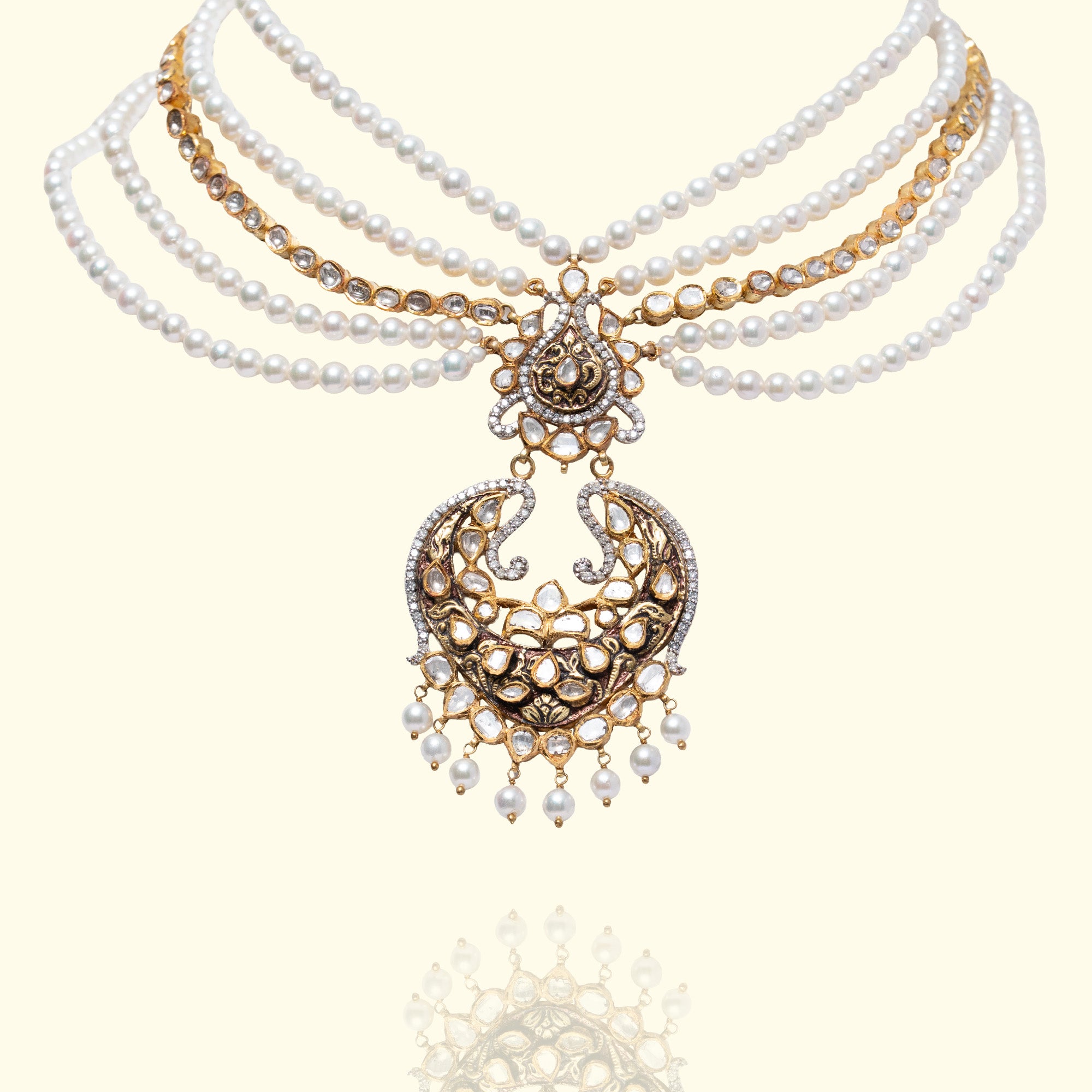 A wide gold choker featuring geometric and floral polki diamond settings, topped with a row of small pearls and finished with a lush fringe of vibrant emerald bead drops.
A multi-strand japanese freshater pearl necklace featuring an ornate gold pendant with intricate polki diamond scrollwork in a traditional crescent (chandbali) silhouette, accented with hanging pearl drops.