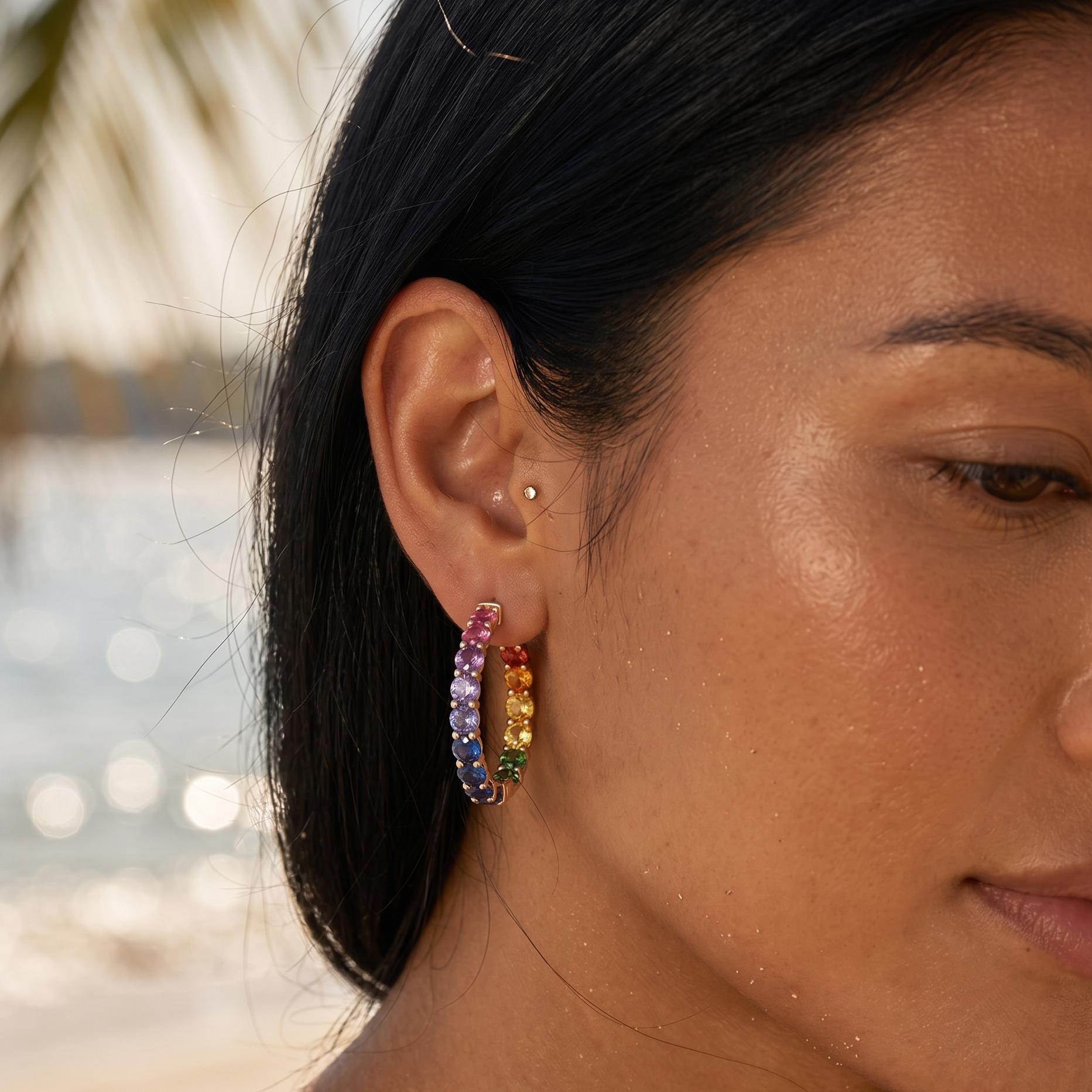 Close-up of a woman's ear wearing colorful rainbow/multisapphire hoop earrings with a blurred beach background, 14kt gold, the alchemy studio, delhi
