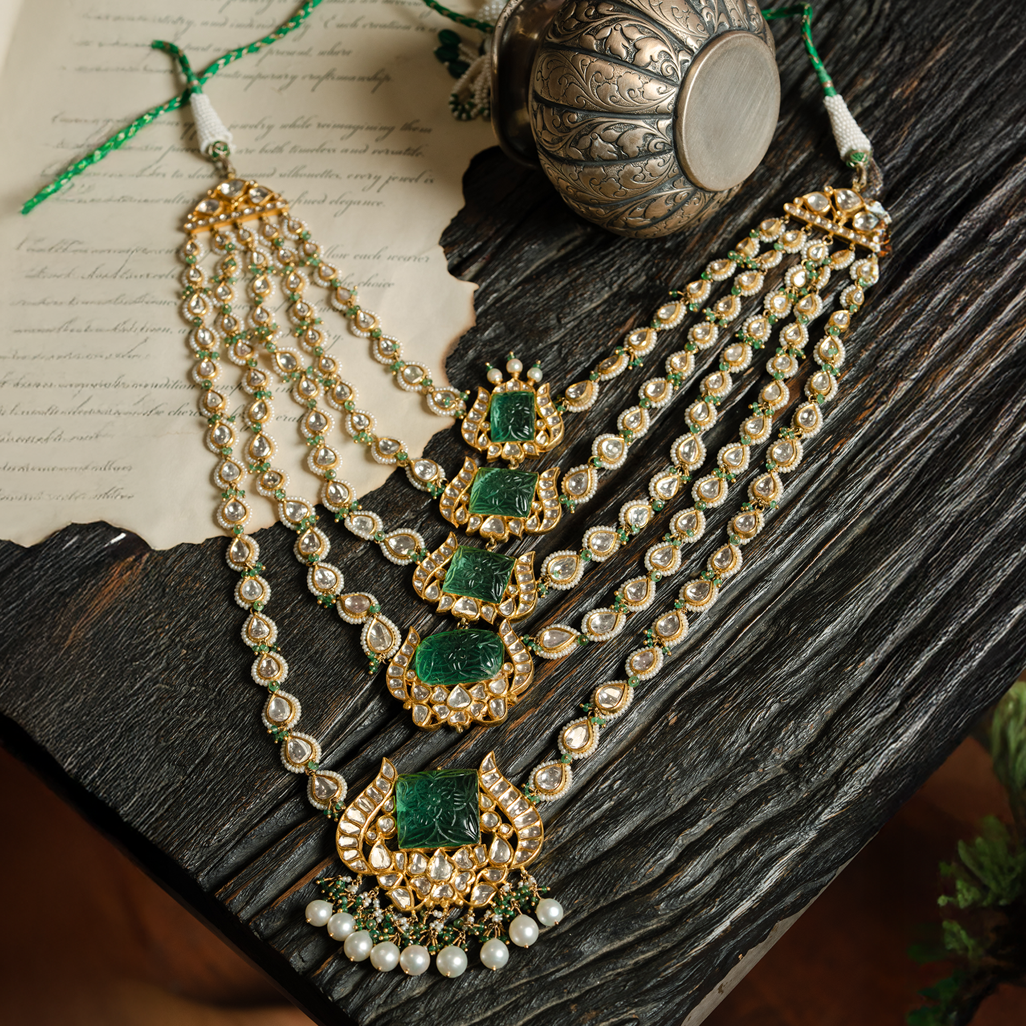 A flat-lay view of a magnificent multi-strand Satlada-style necklace showcasing five graduated carved emerald plaques set in ornate gold floral frames, with strands of teardrop polki diamonds and a fringe of pearls and emerald beads.