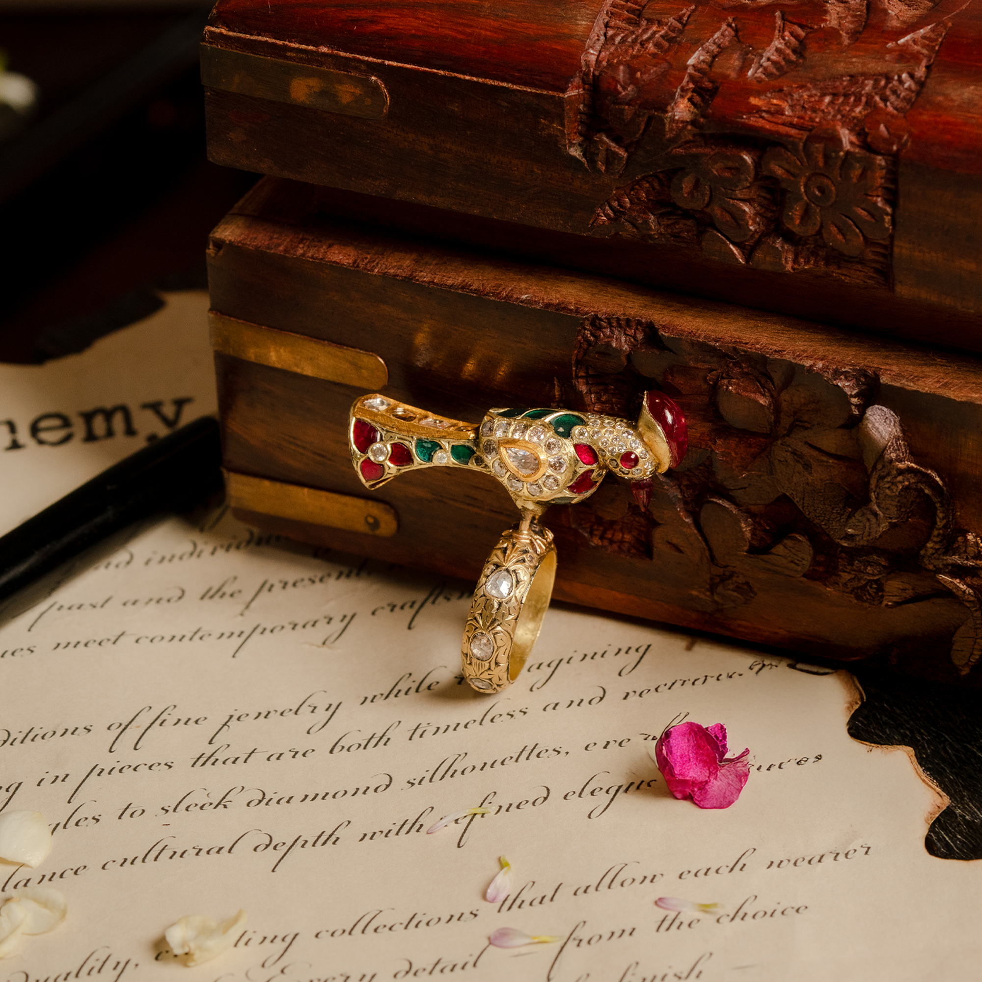 Close-up of a gold bird-wrap ring with uncut polki diamond, rubies and emeralds, styled on vintage parchment next to an antique wooden box.