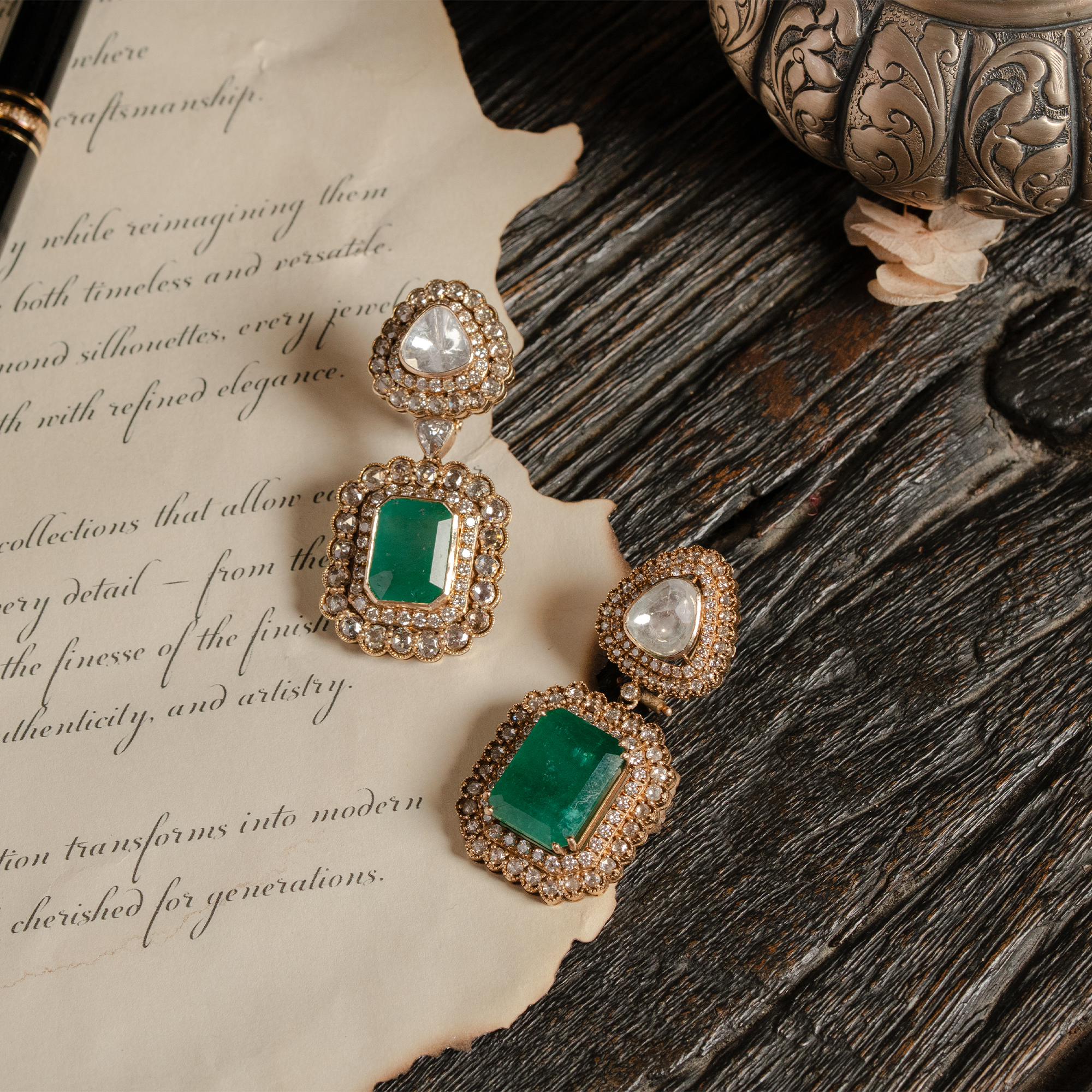 Pair of gold Polki and emerald drop earrings featuring an emerald-cut central emerald surrounded by a diamond halo, styled on vintage parchment with an antique silver box.