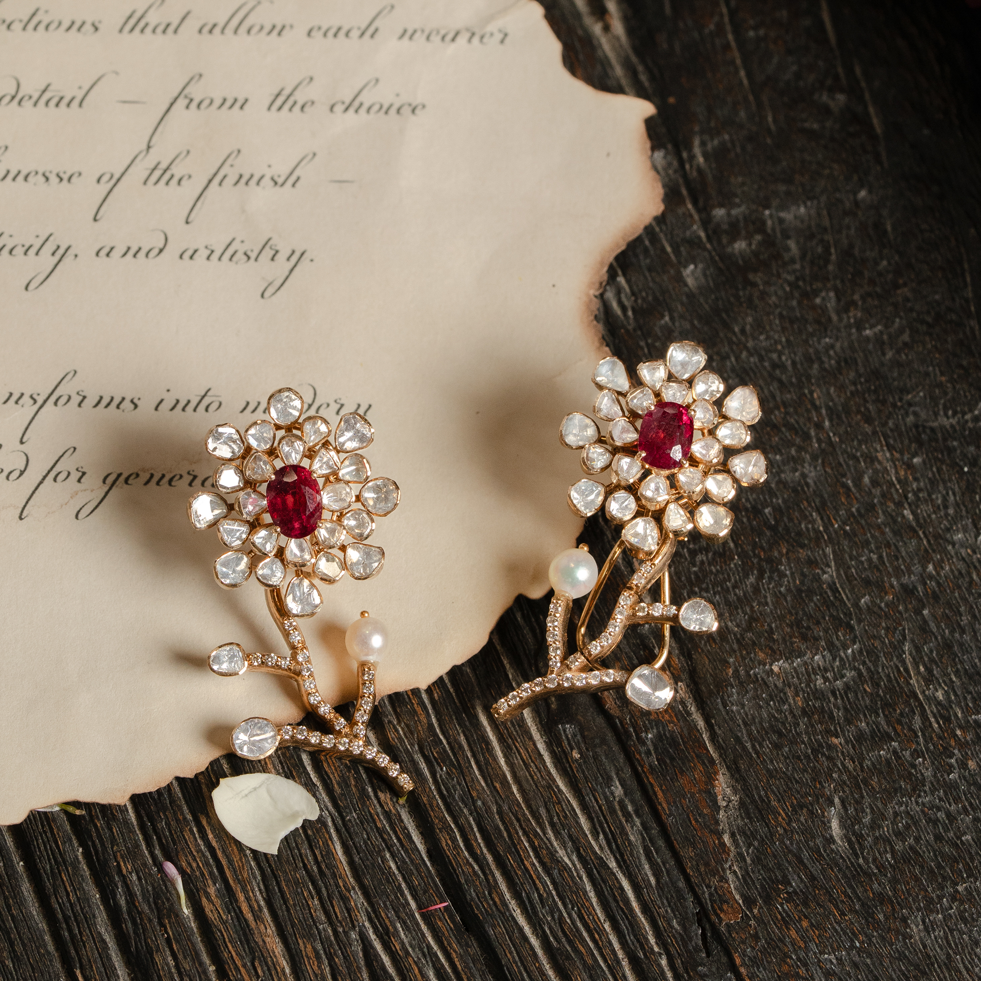 A pair of artistic gold polki ruby earrings shaped like delicate blooming flowers, featuring a central oval-cut ruby surrounded by a petal-like halo of uncut diamonds, set atop a curved stem accented with pavé diamonds and a single seed pearl.