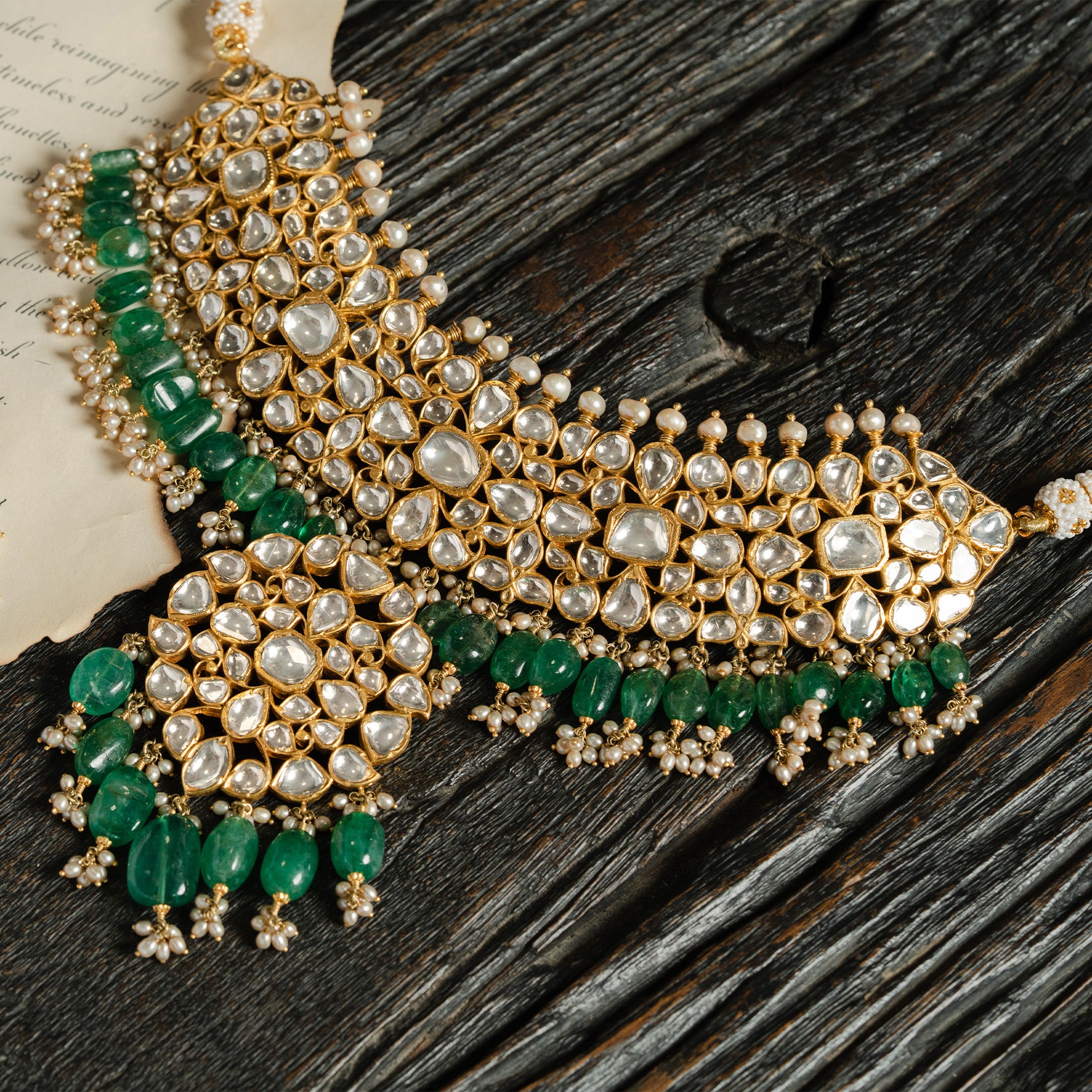 A high-neck gold polki choker featuring a dense mosaic of pear and heart-shaped uncut diamonds, accented with a double fringe of vibrant emerald beads and a central, oversized polki diamond pendant.