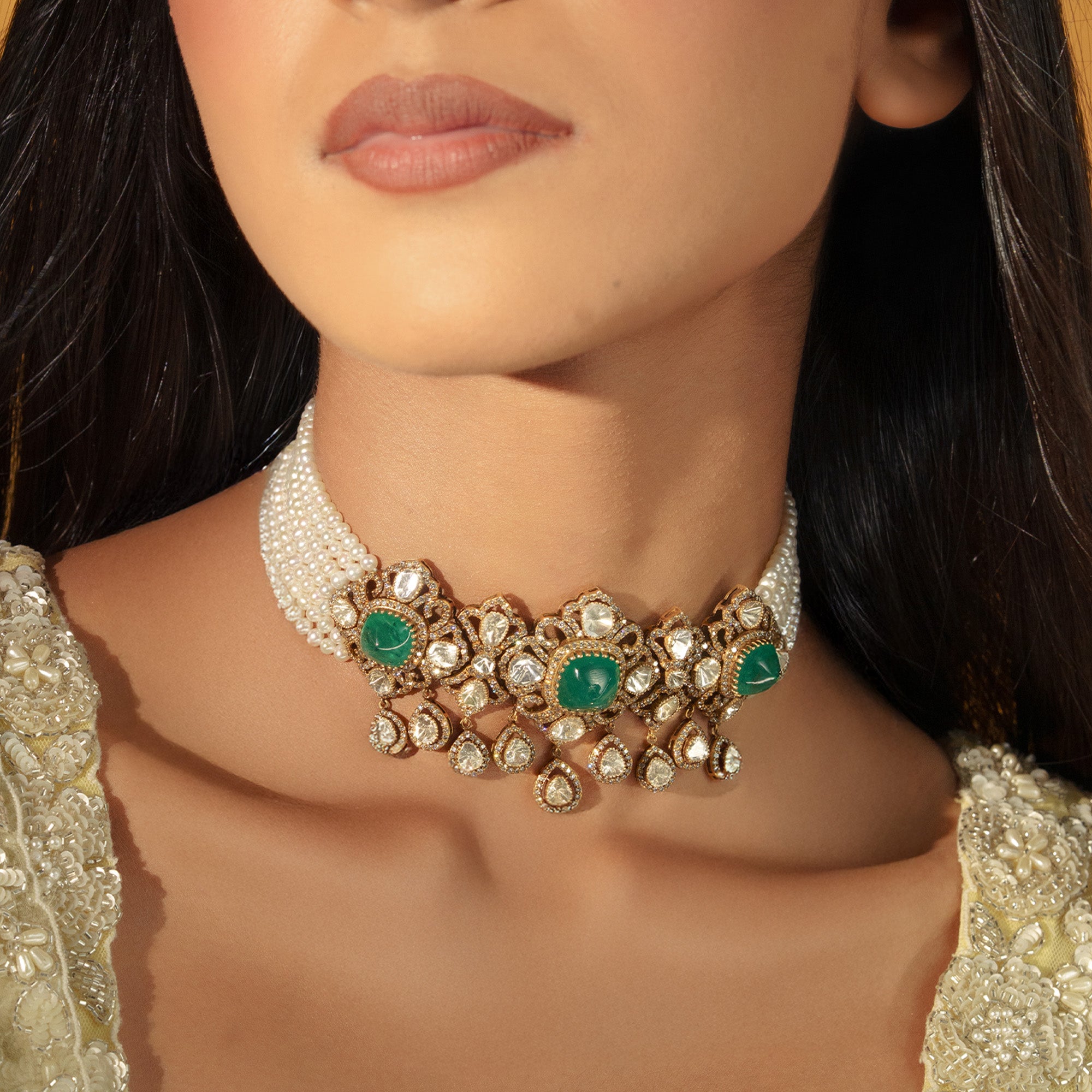 A model wearing a regal choker featuring multiple strands of creamy seed japanese freshwater pearls and an ornate gold centerpiece set with three large emerald cabochons, surrounded by intricate polki diamond scrollwork and hanging pear-shaped uncut diamond drops.
