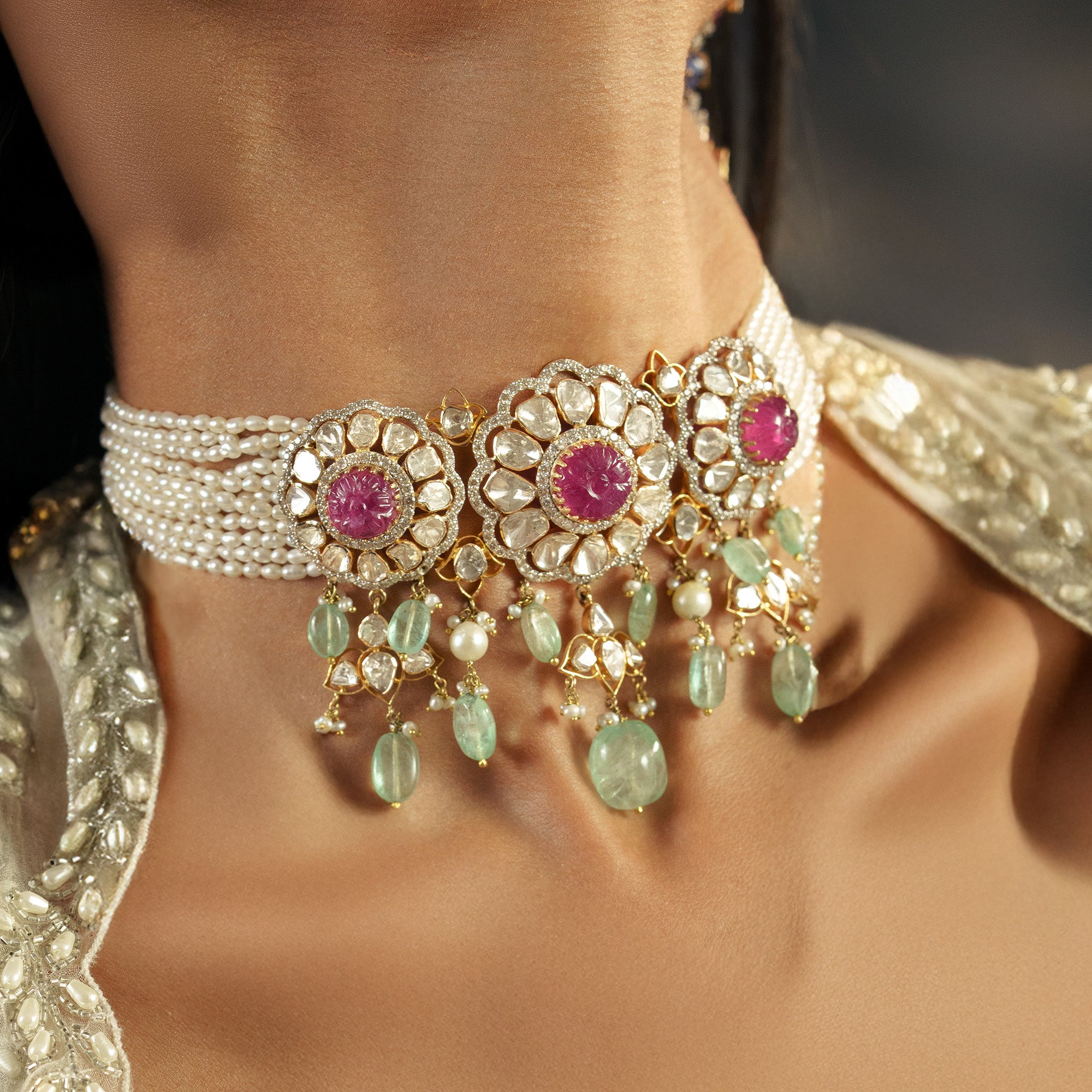 A model wearing a multi-strand japanese freshwater pearl choker featuring three large, circular polki diamond medallions with carved ruby cabochon centers, finished with a delicate fringe of mint-green emerald drops and seed pearls.