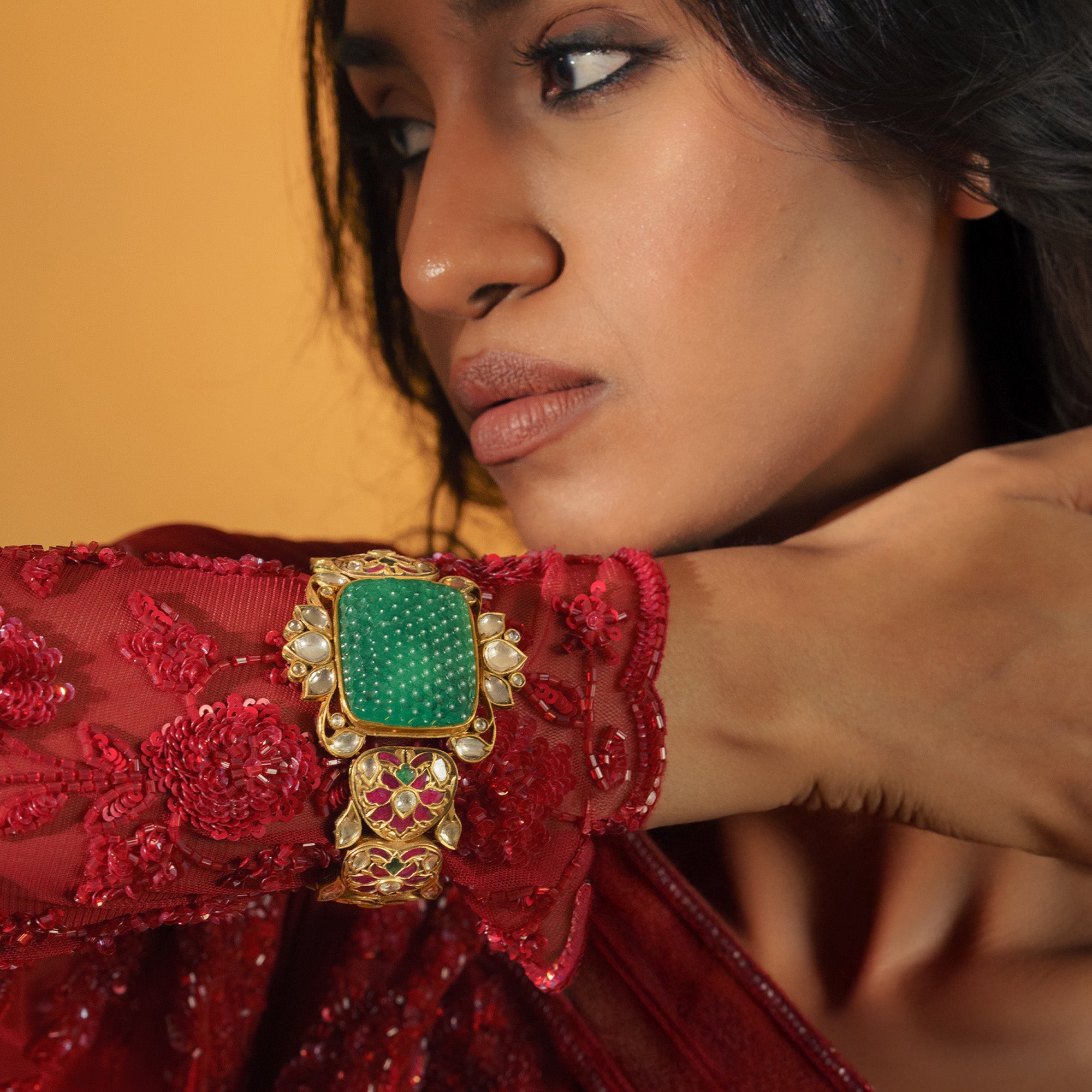 Editorial close-up of a model wearing a large carved emerald, polki, ruby cuff bracelet and a bird-motif ring, paired with a red sequined ethnic outfit.