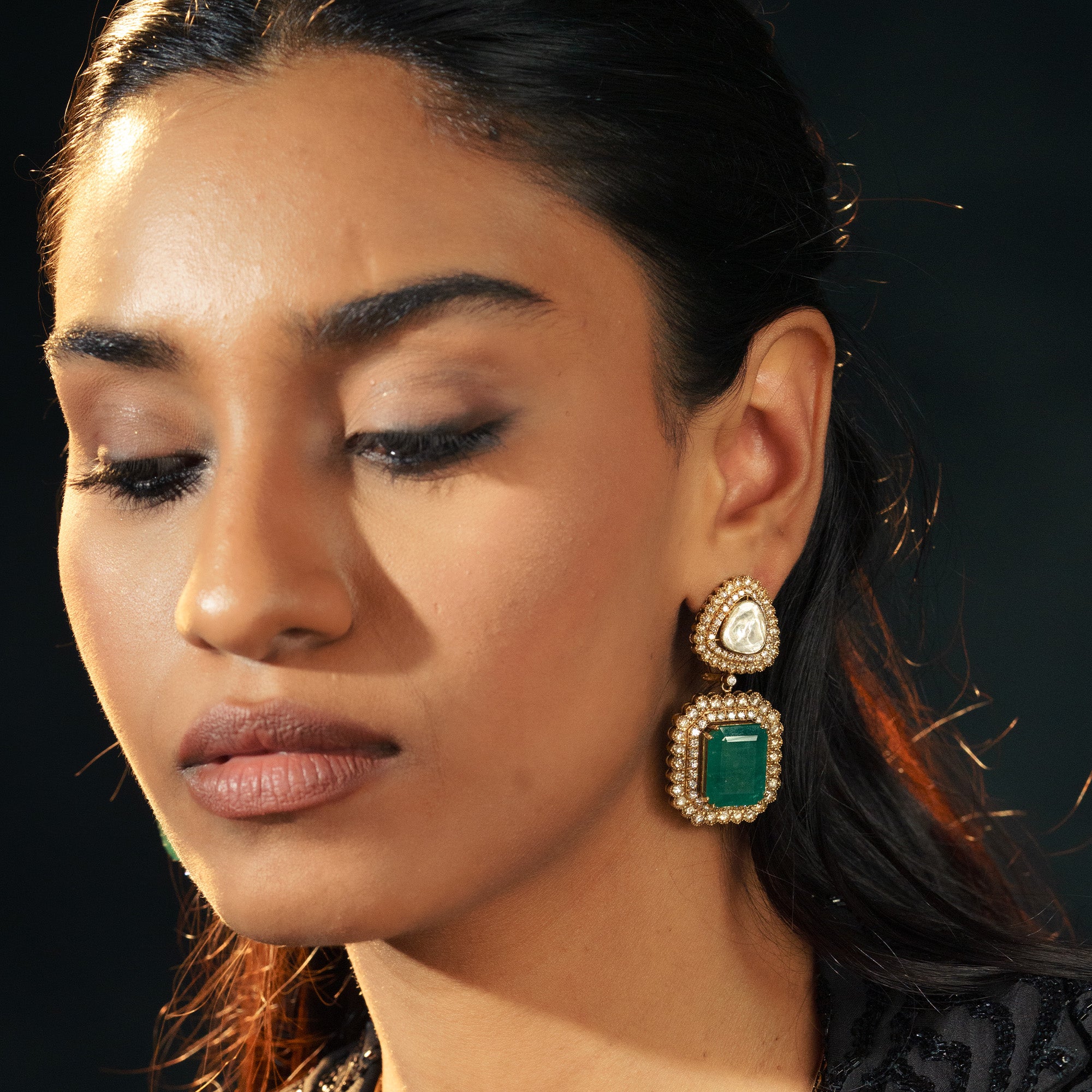 A model wearing elegant double-drop earrings featuring a pear-shaped polki diamond stud and a large, rectangular emerald-cut emerald, both framed by shimmering pavé diamond halos.