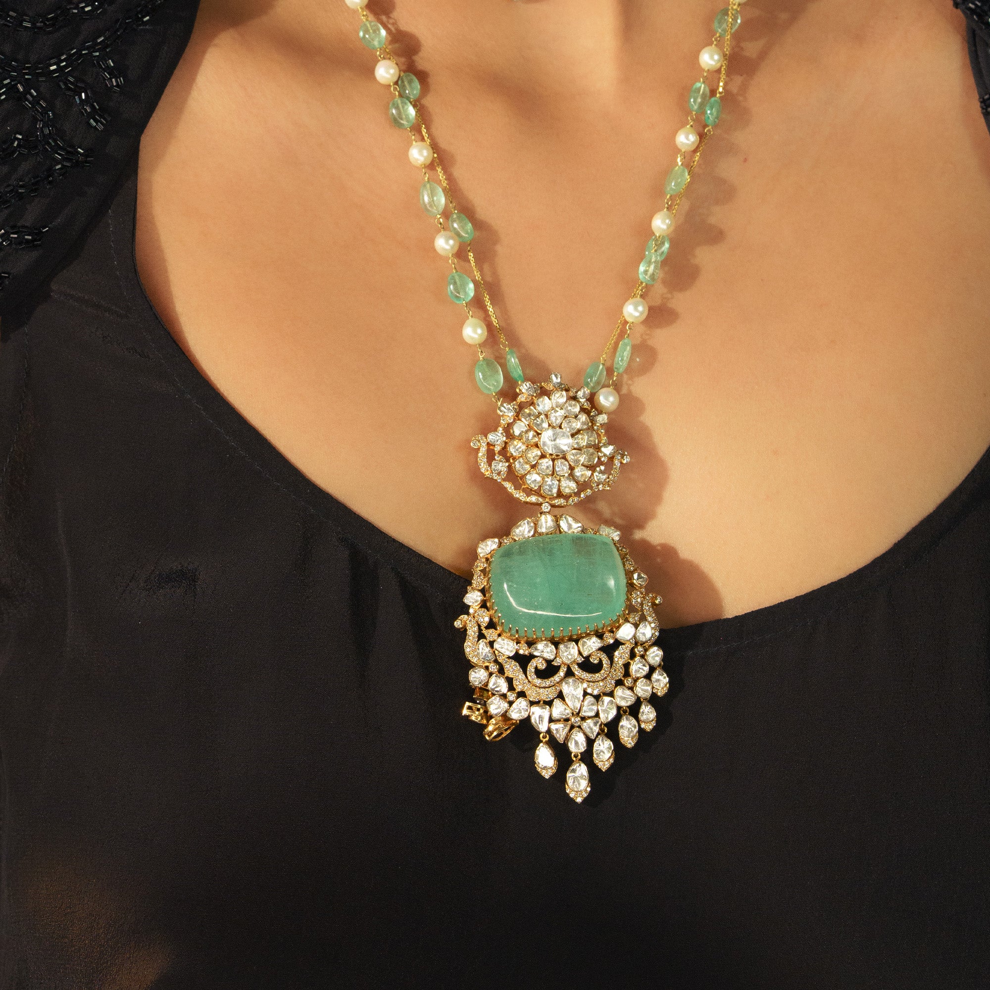 A model is wearing a regal gold necklace featuring a delicate chain of emerald beads and pearls, leadings to a massive, rectangular emerald cabochon pendant framed by ornate polki diamond scrollwork and cascading pear-shaped diamond drops.