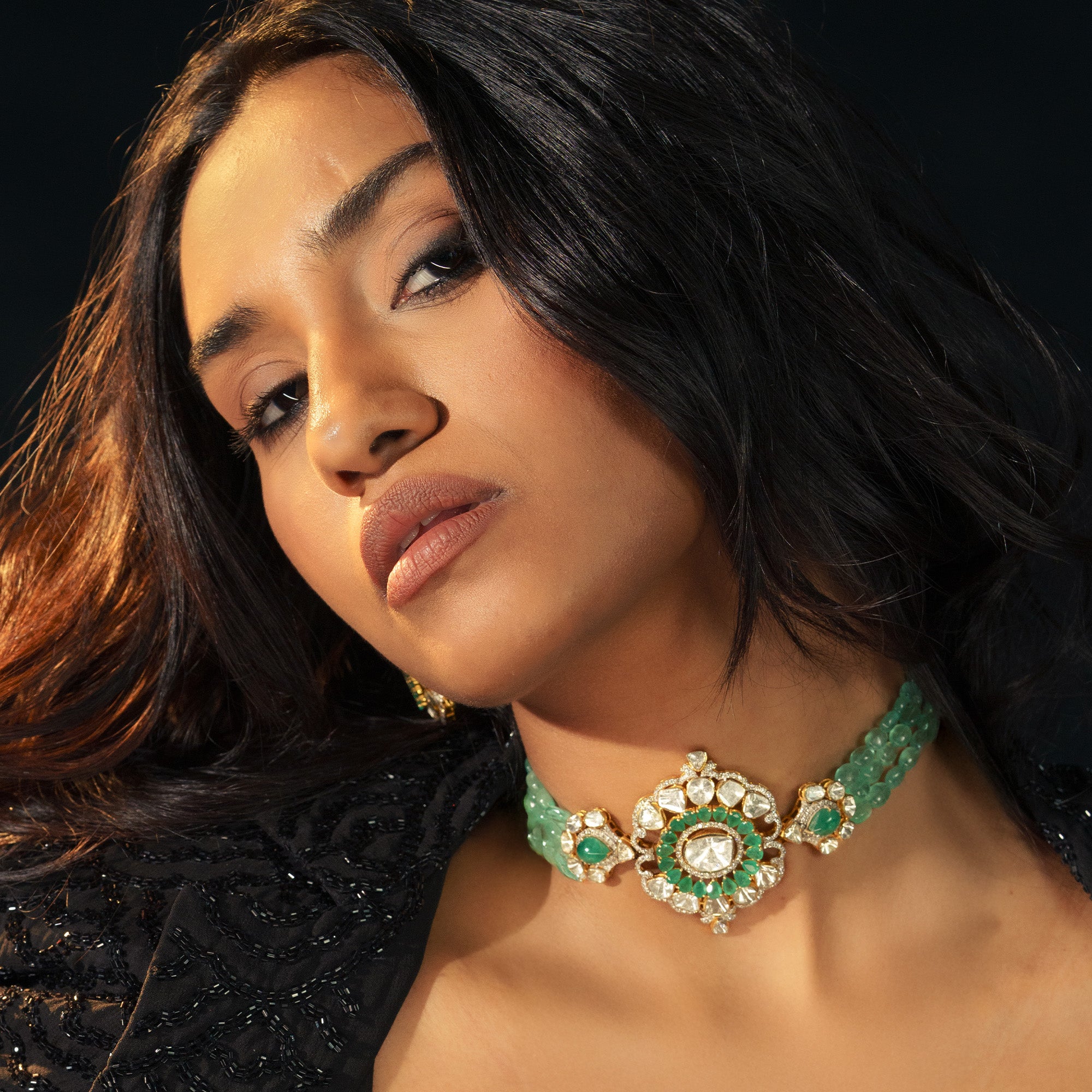 A model wearing a regal choker featuring three strands of vibrant emerald beads and a large, ornate central pendant with a sunburst of polki diamonds and a halo of pear-shaped emerald.