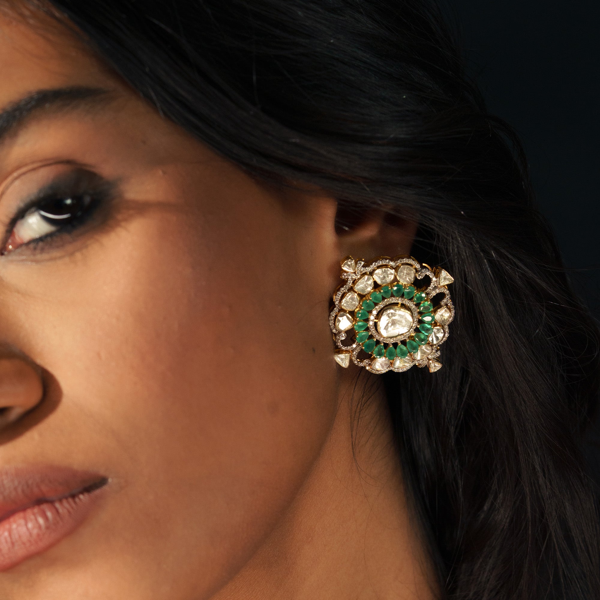 A model wearing an oversized, ornate polki diamond stud earring featuring a central polki diamond surrounded by a halo of vibrant emeralds and an outer frame of larger uncut diamond stones.