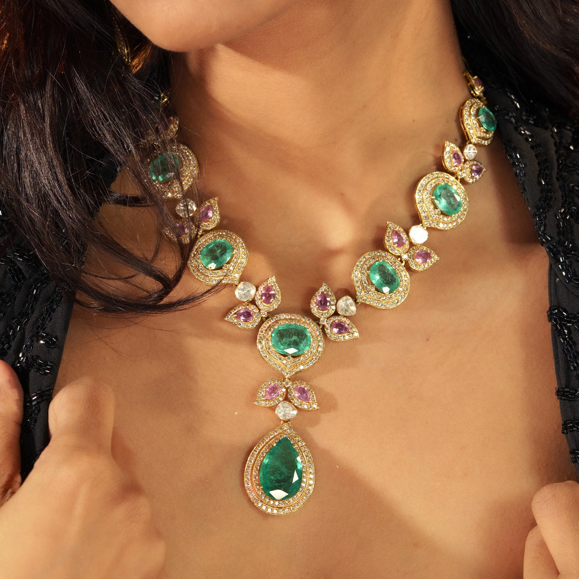 A model wearing a sophisticated gold polki emerald tourmaline necklace featuring oval emerald set within diamond-encrusted circular frames, linked by pink tourmaline and polki diamond butterfly motifs, and finished with a large, pear-shaped emerald drop.