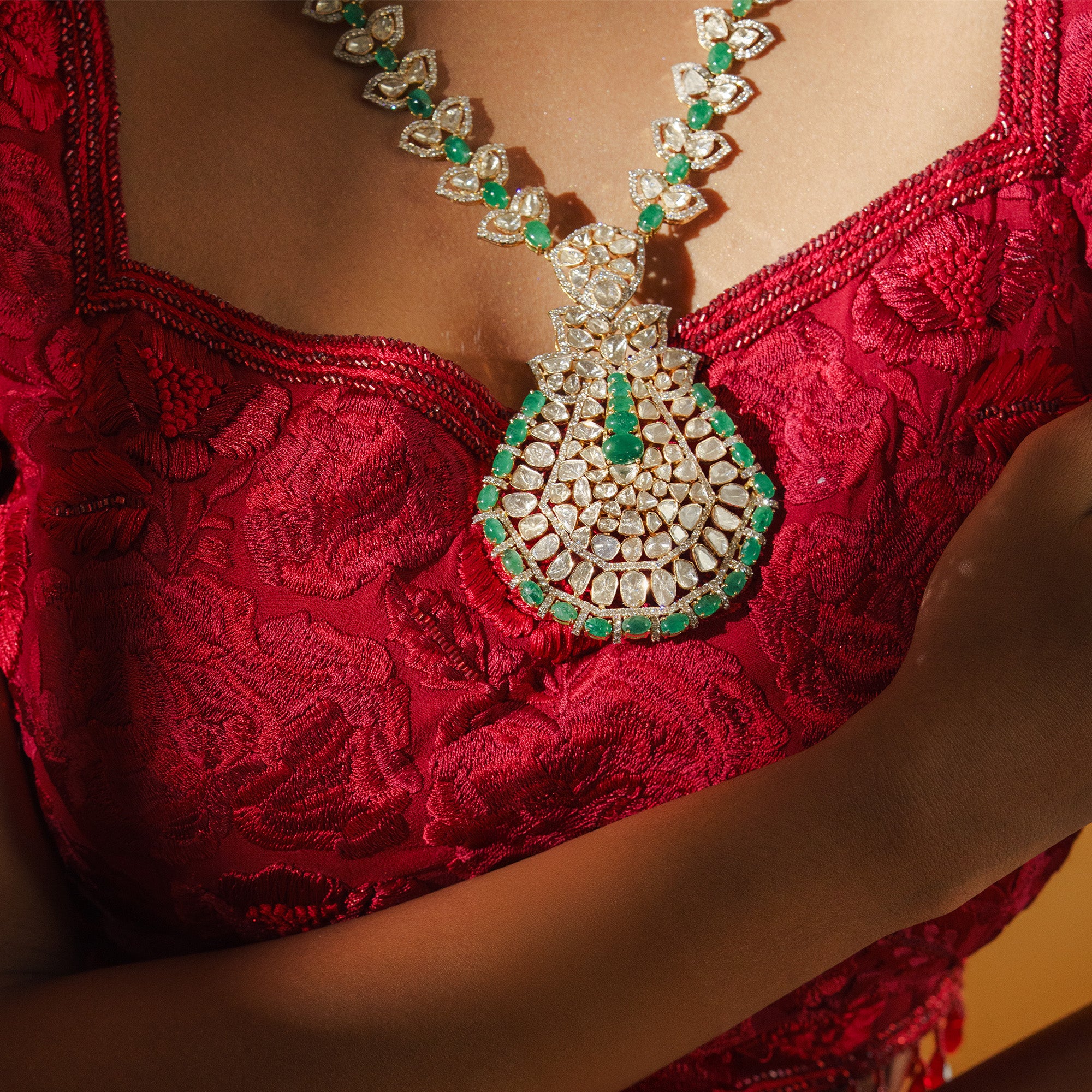 Close-up of a model wearing a heavy Polki diamond pendant necklace with an intricate teardrop design, edged with vibrant green emerald cabochons against a red embroidered dress.
