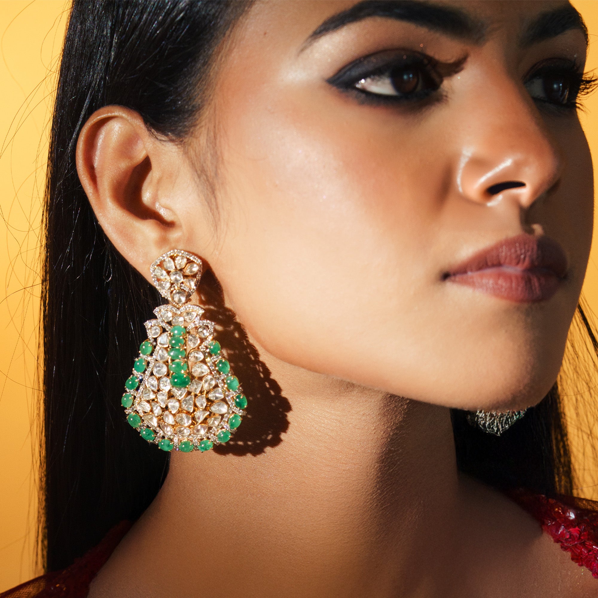 A model wearing a large gold Polki statement earrings featuring a complex lattice of uncut diamonds and a vertical row of emerald cabochons with a vibrant border of emerald-bead fringe.