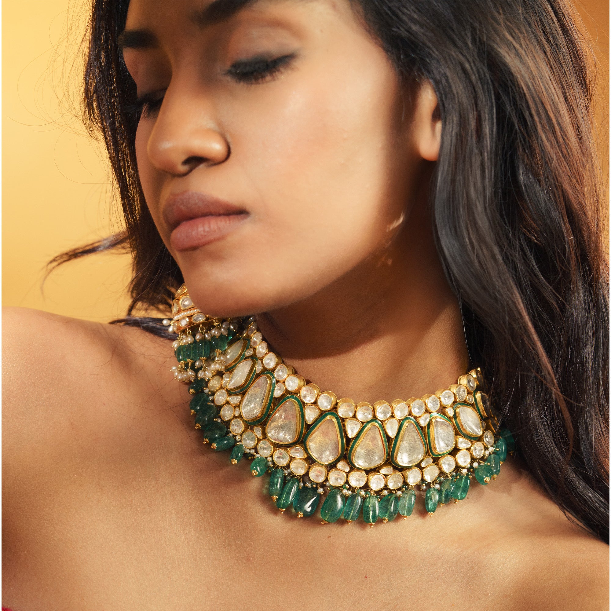 A model wearing a wide, statement gold choker featuring large, teardrop-shaped polki diamonds rimmed with green enamel, finished with a lush fringe of vibrant emerald beads and small pearls.