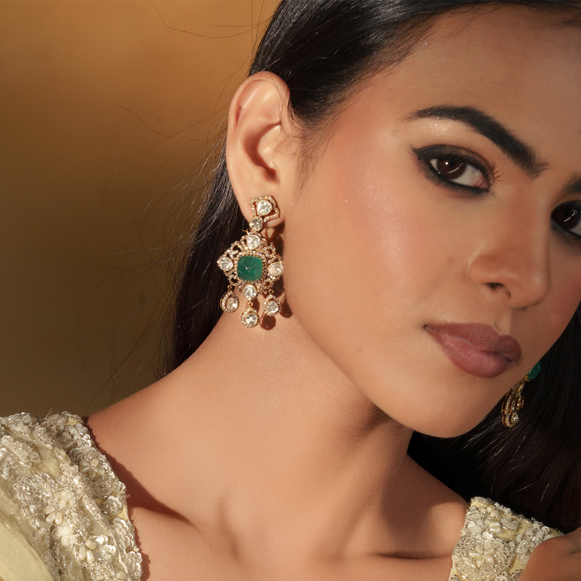 Close-up of a model wearing gold Polki earrings with a central green emerald cabochon surrounded by an intricate diamond lattice and dangling Polki charms in india