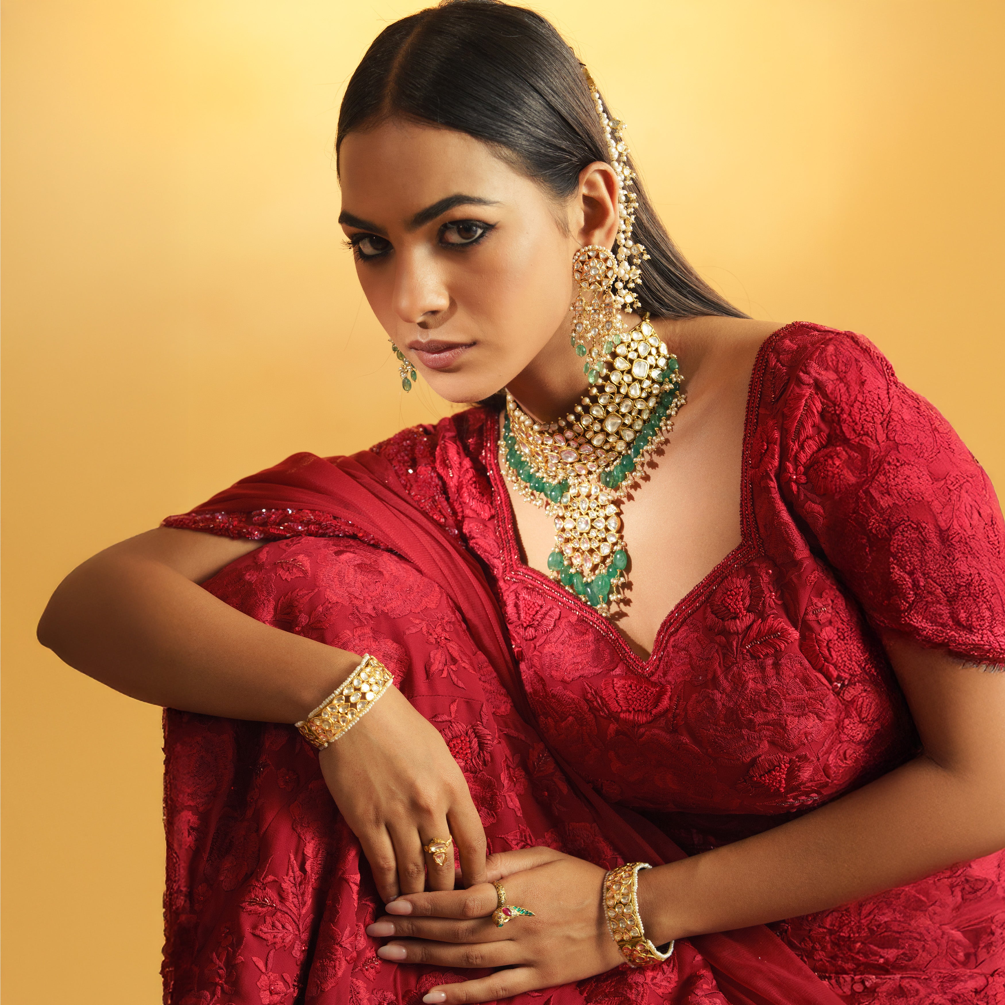 A model in a red embroidered ensemble wearing a heavy tiered polki diamond necklace with a lush fringe of vibrant emerald beads, paired with oversized emerald chandelier earrings and matching polki style bangles paired with bird motif emerald uncut diamond ring.